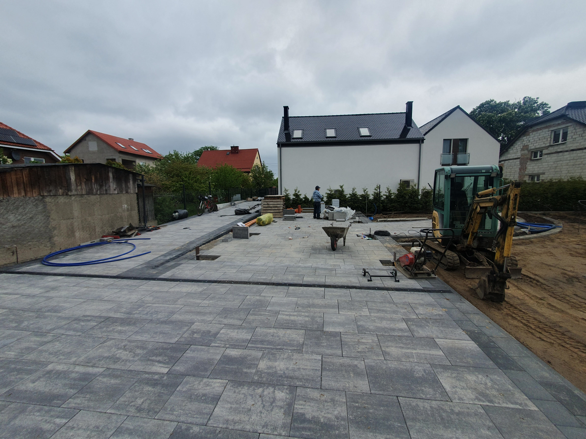 Układanie szarej kostki brukowej na posesji domu jednorodzinnego, widoczny minikoparka, częściowo ułożona nawierzchnia, przygotowane materiały i narzędzia.