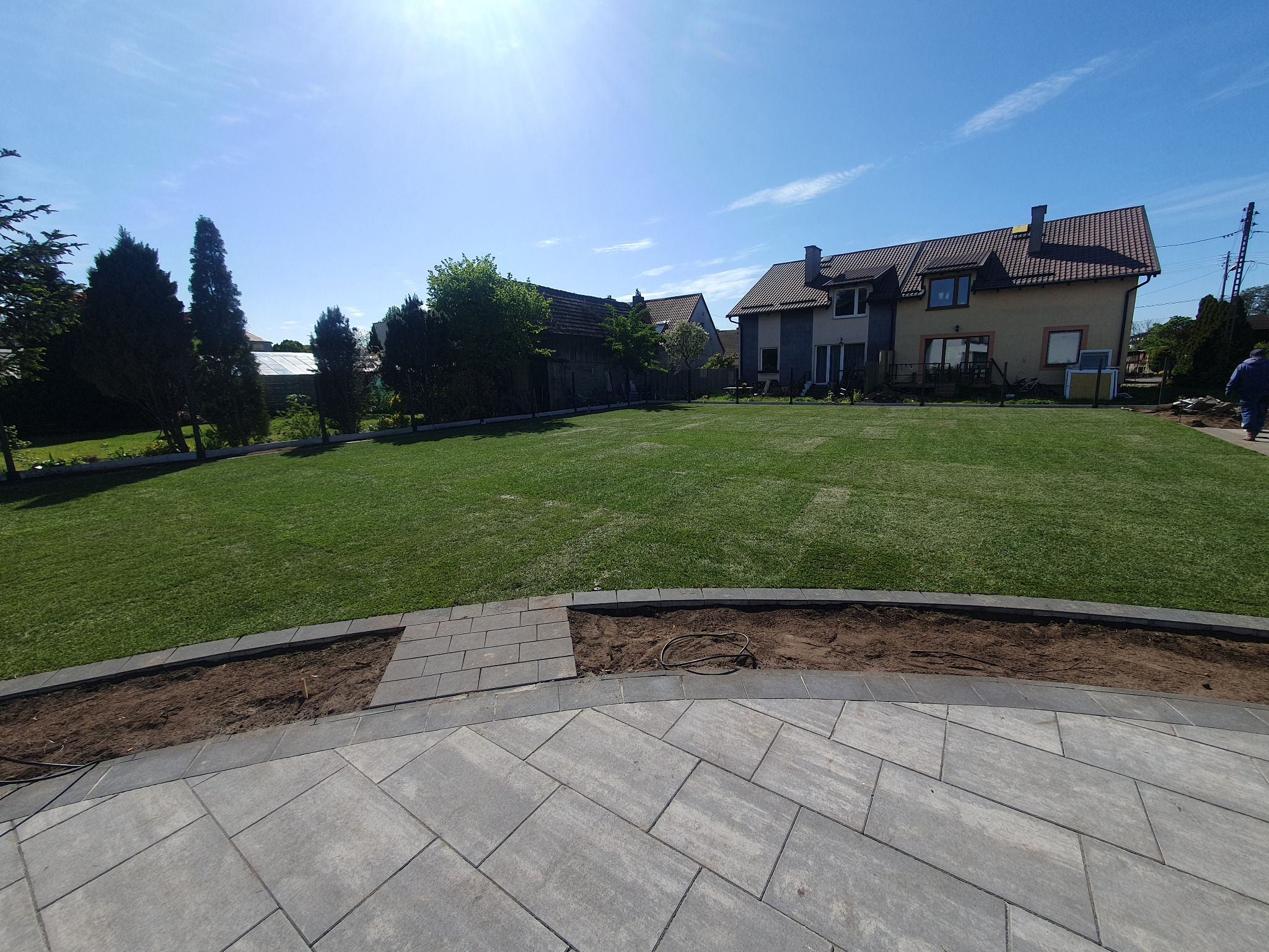 Nowoczesna kostka brukowa w szarym odcieniu tworzy taras z obrzeżami. W tle zielony trawnik i domy. Częściowo niezakończone rabaty kwiatowe.