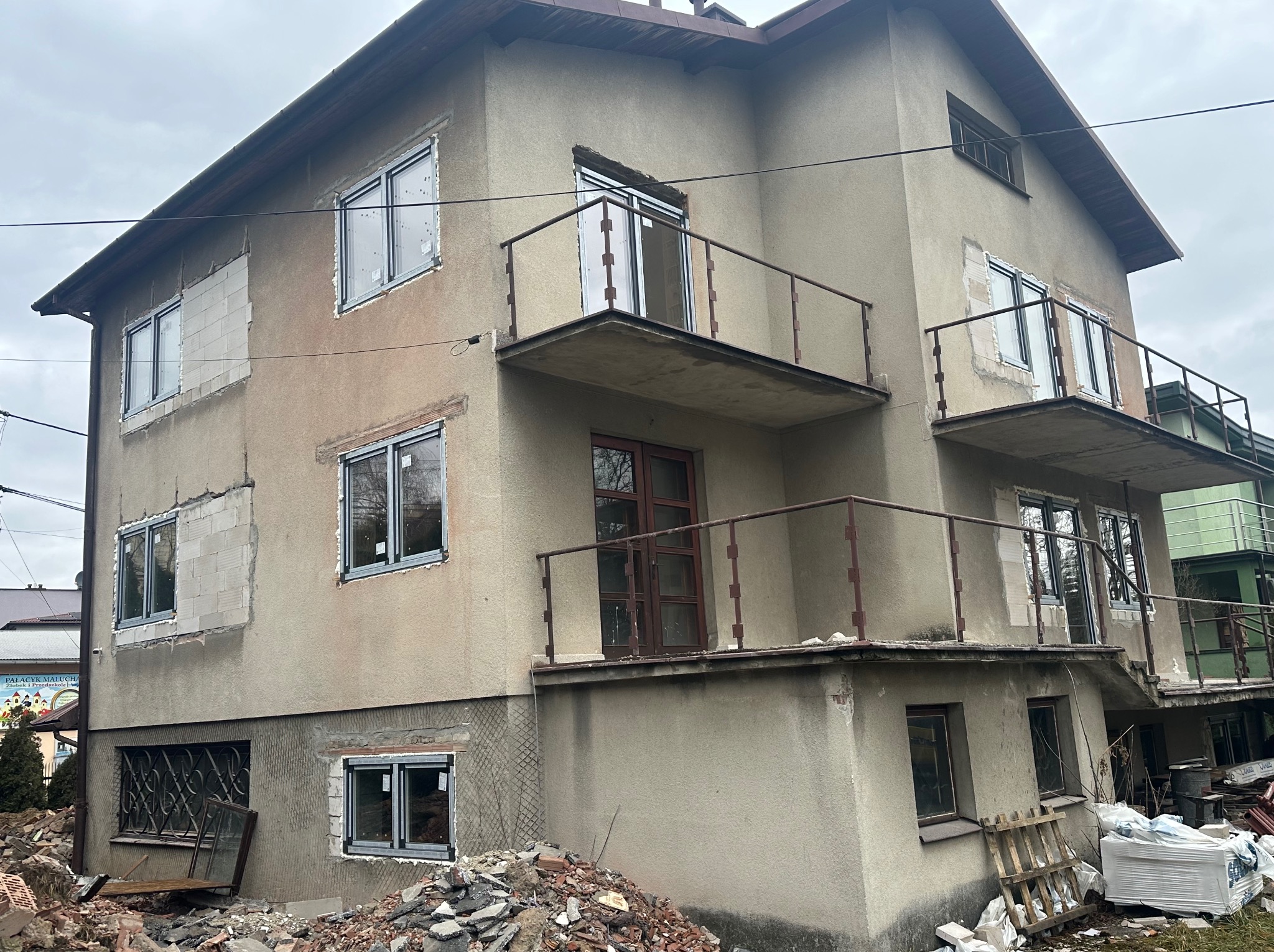 Dom w trakcie wymiany okien, widoczne nowe okna PCV w surowych otworach, gruz i materiały budowlane na zewnątrz. Częściowo ocieplona ściana budynku.
