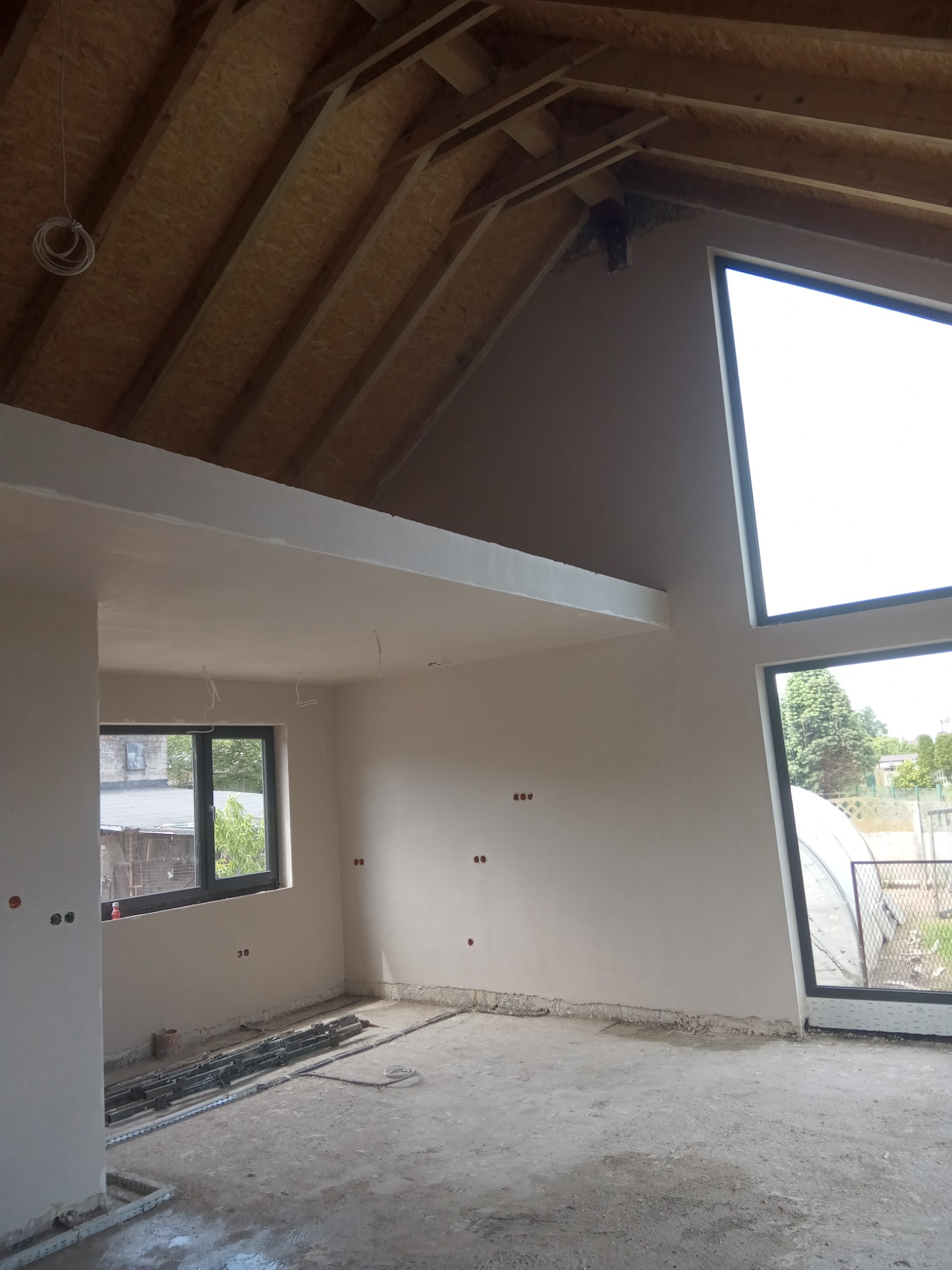 Wnętrze po tynkowaniu ścian w Tarnowskim Młynie. Widoczne gładkie, białe ściany, surowy strop i duże okna. Na podłodze widoczne materiały budowlane.