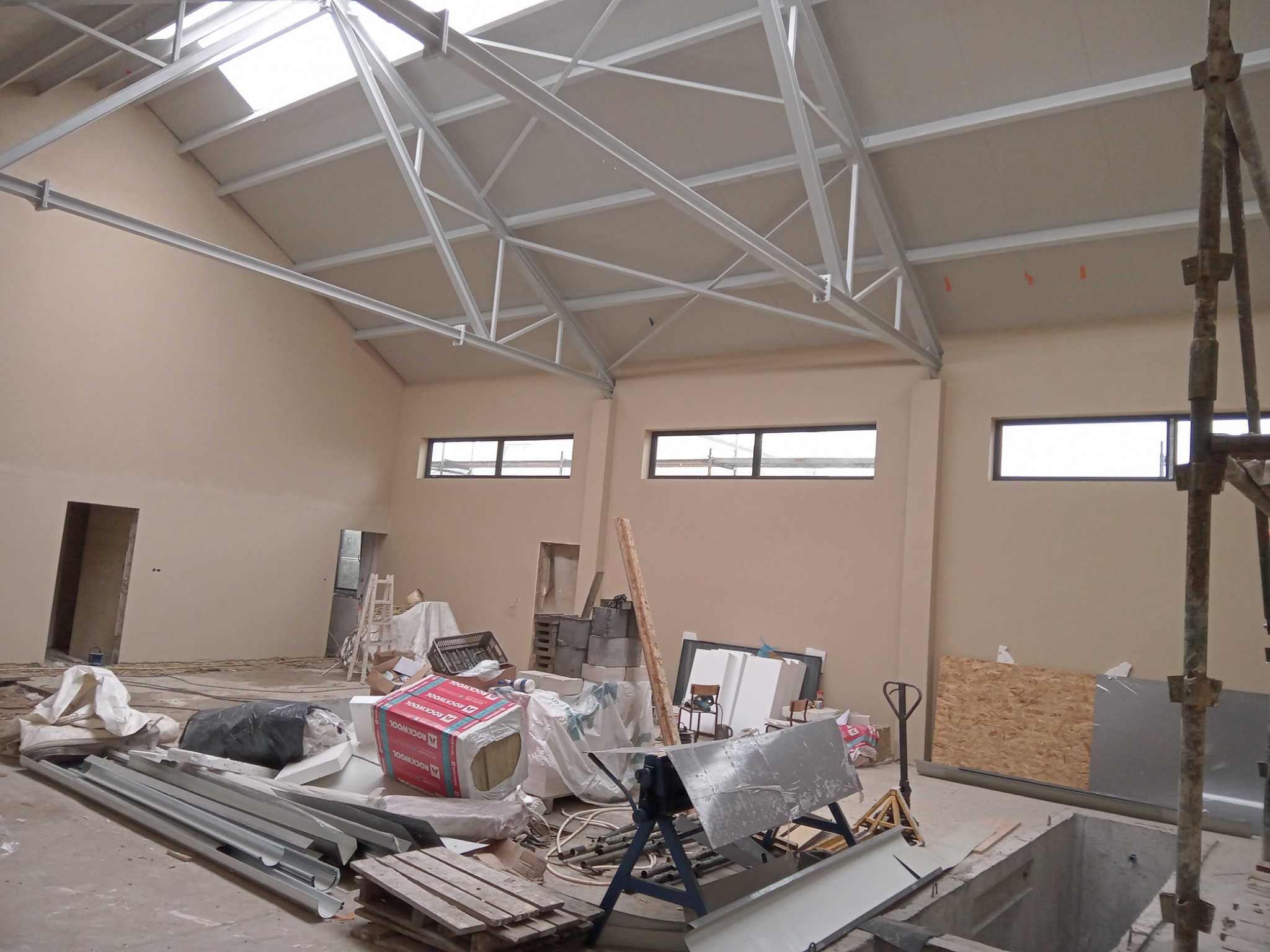 Wnętrze budynku w trakcie tynkowania: widoczne rusztowania, okna, konstrukcja dachu i materiały budowlane. Ściany w jasnym odcieniu beżu, przygotowane do dalszych prac wykończeniowych.