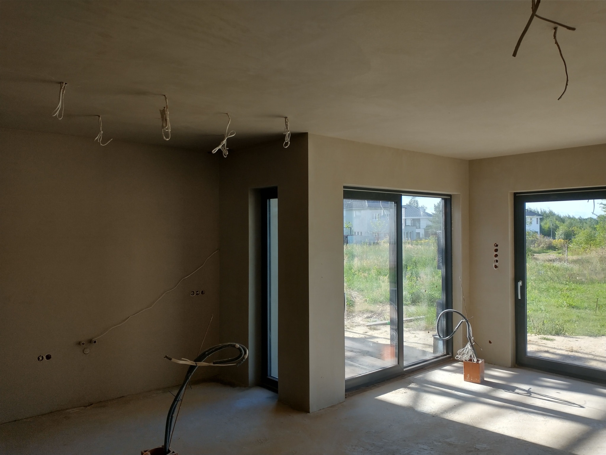 Wnętrze domu po tynkowaniu: gładkie ściany w odcieniach beżu, widoczne kable instalacji elektrycznej, duże okna przesuwne z widokiem na zieleń. Na podłodze resztki materiałów.