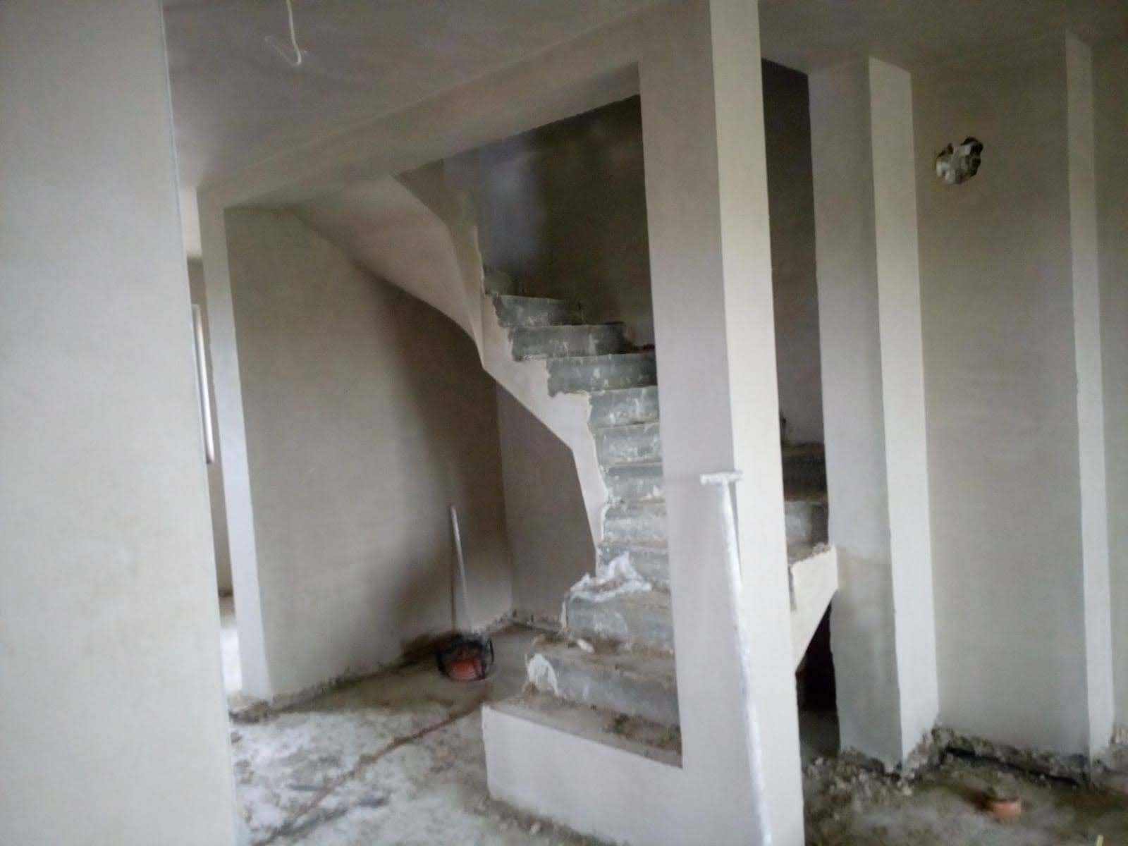 Wnętrze w trakcie tynkowania: ściany, schody w szarym tynku. Widoczne niedoskonałości i narzędzia. Neutralne oświetlenie, surowy stan wykończenia.