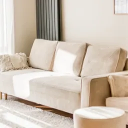 Jasny salon z beżową sofą, poduszkami i pufą. Minimalistyczny design, miękki dywan. Lekkie, naturalne światło przez okno z firanką. Widok zza framugi drzwi.