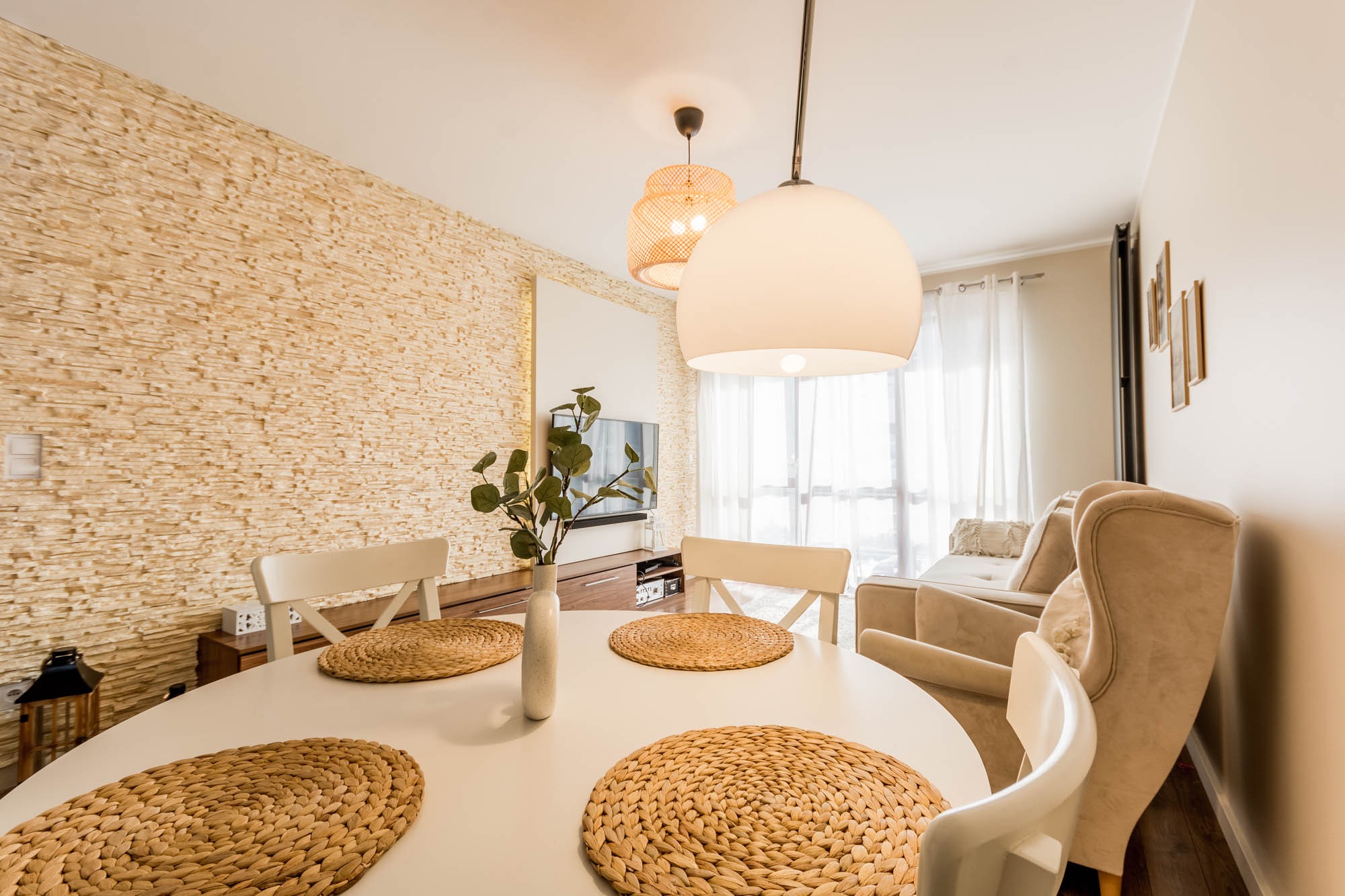 Jasna jadalnia z okrągłym stołem, plecionymi podkładkami i dekoracyjną ścianą z kamienia. Dwie lampy wiszące nad stołem. W tle salon z beżową sofą i telewizorem.