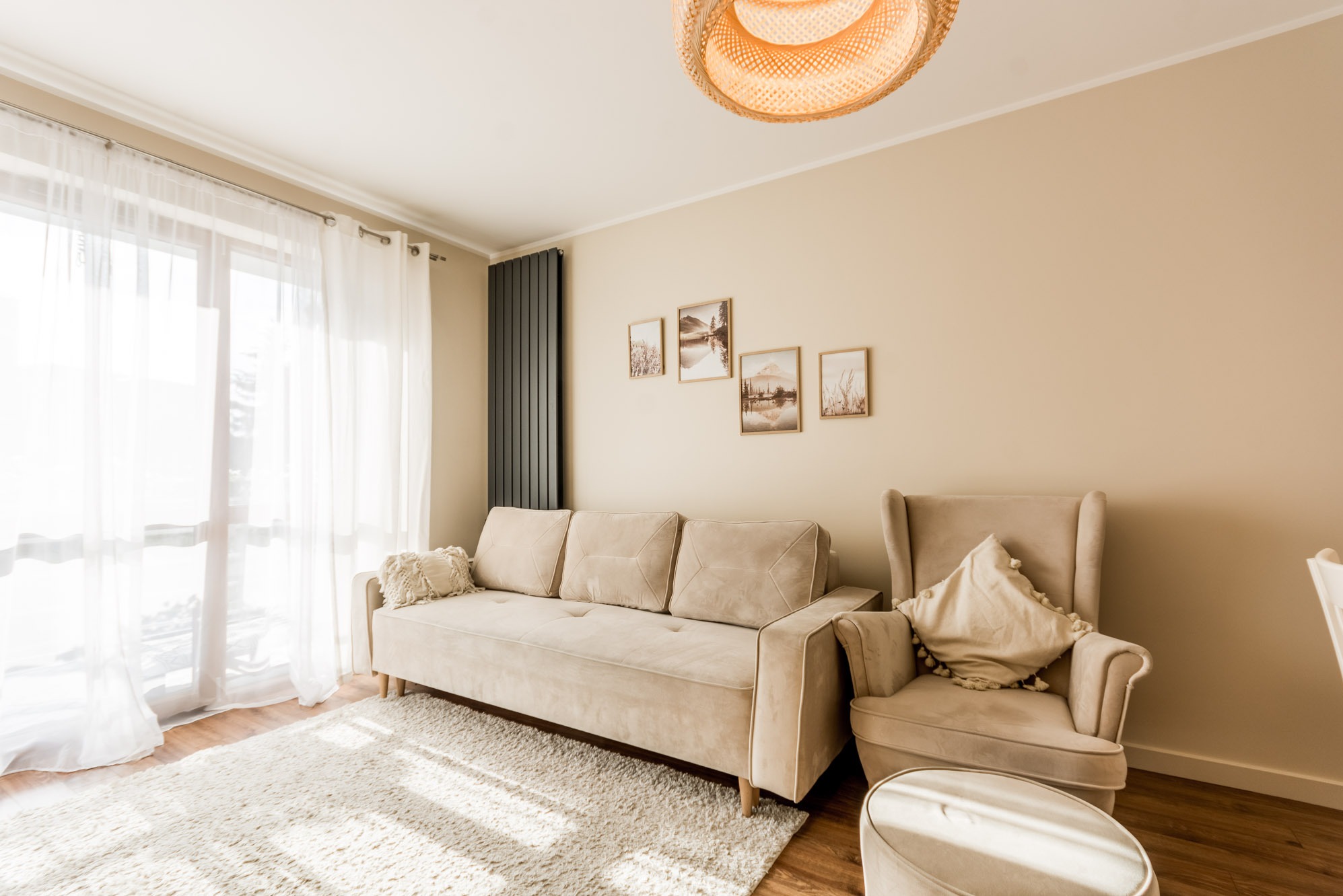 Jasny salon z beżową sofą, fotelem i puszystym dywanem. Na ścianie grafiki w ramkach, a nad sofą nowoczesny grzejnik. Całość doświetlona naturalnym światłem.