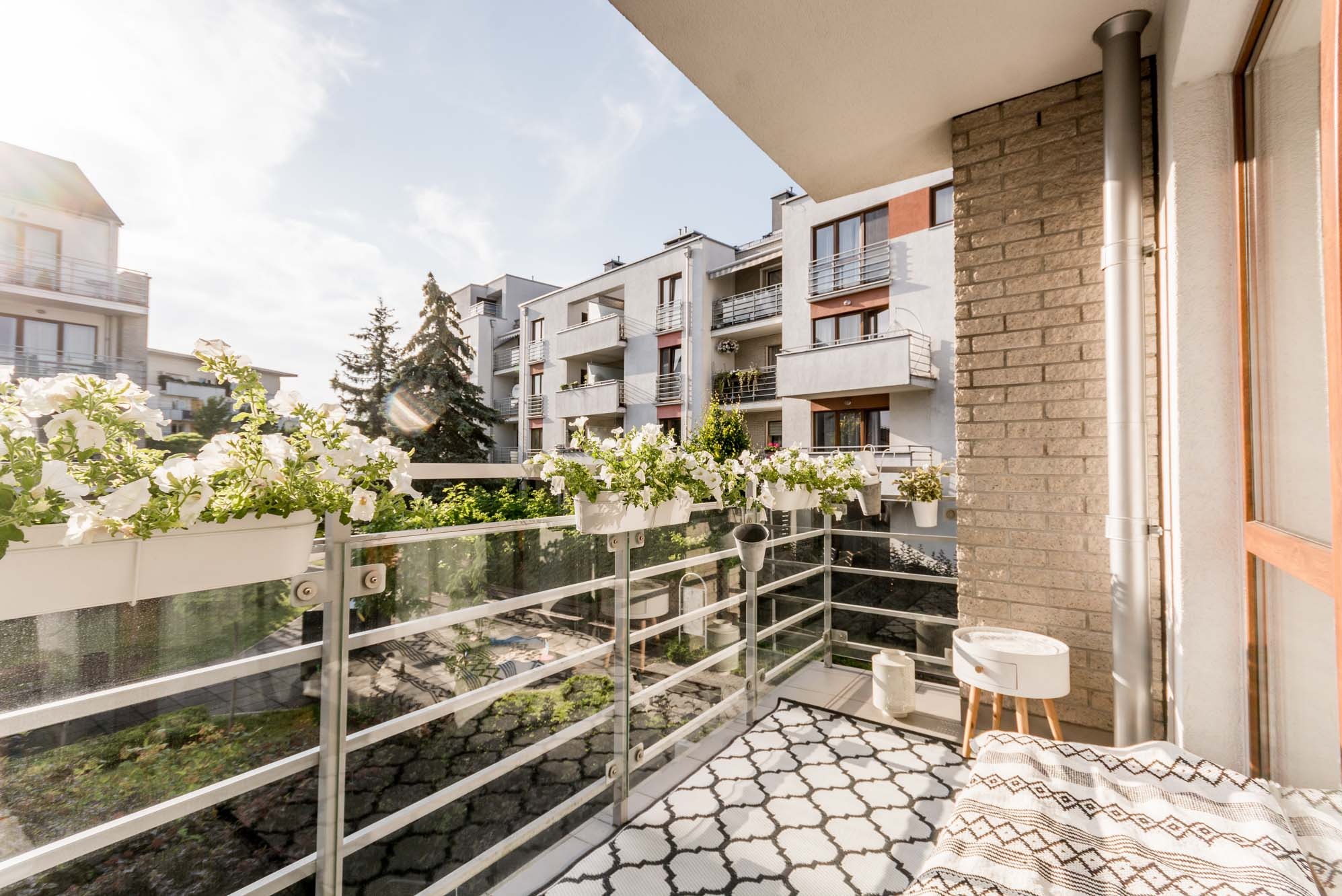 Balkon z białymi kwiatami w donicach, dywanem w geometryczny wzór i widokiem na budynki w tle, oświetlony słonecznym światłem.