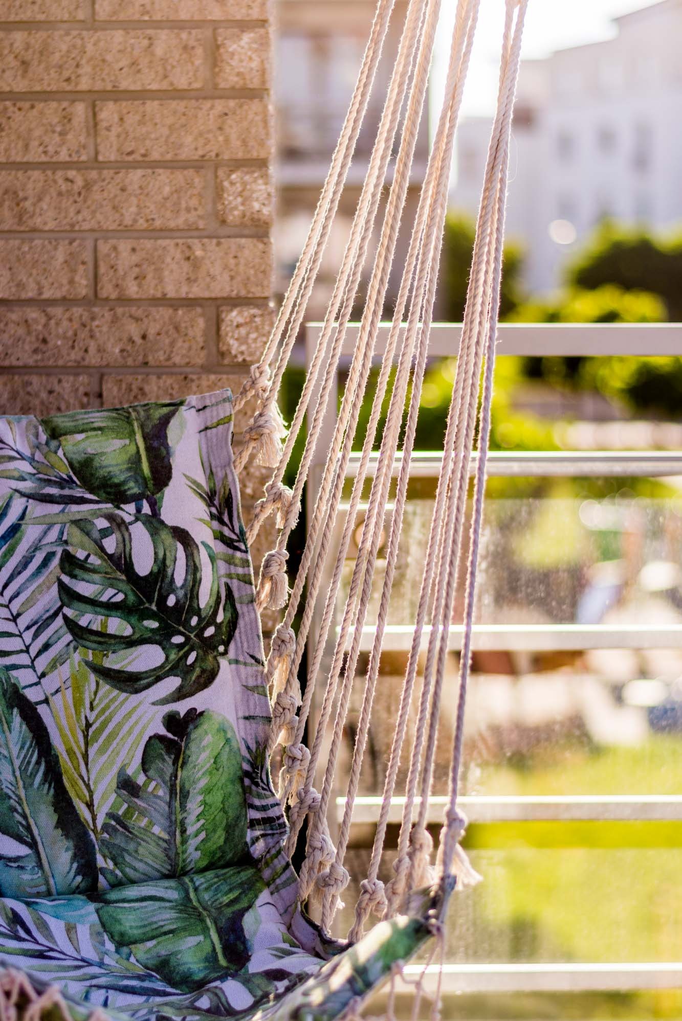 Makro ujęcie wiszącego fotela hamakowego z motywem roślinnym na balkonie w słoneczny dzień. Widoczne sznurki i ceglana ściana w tle.