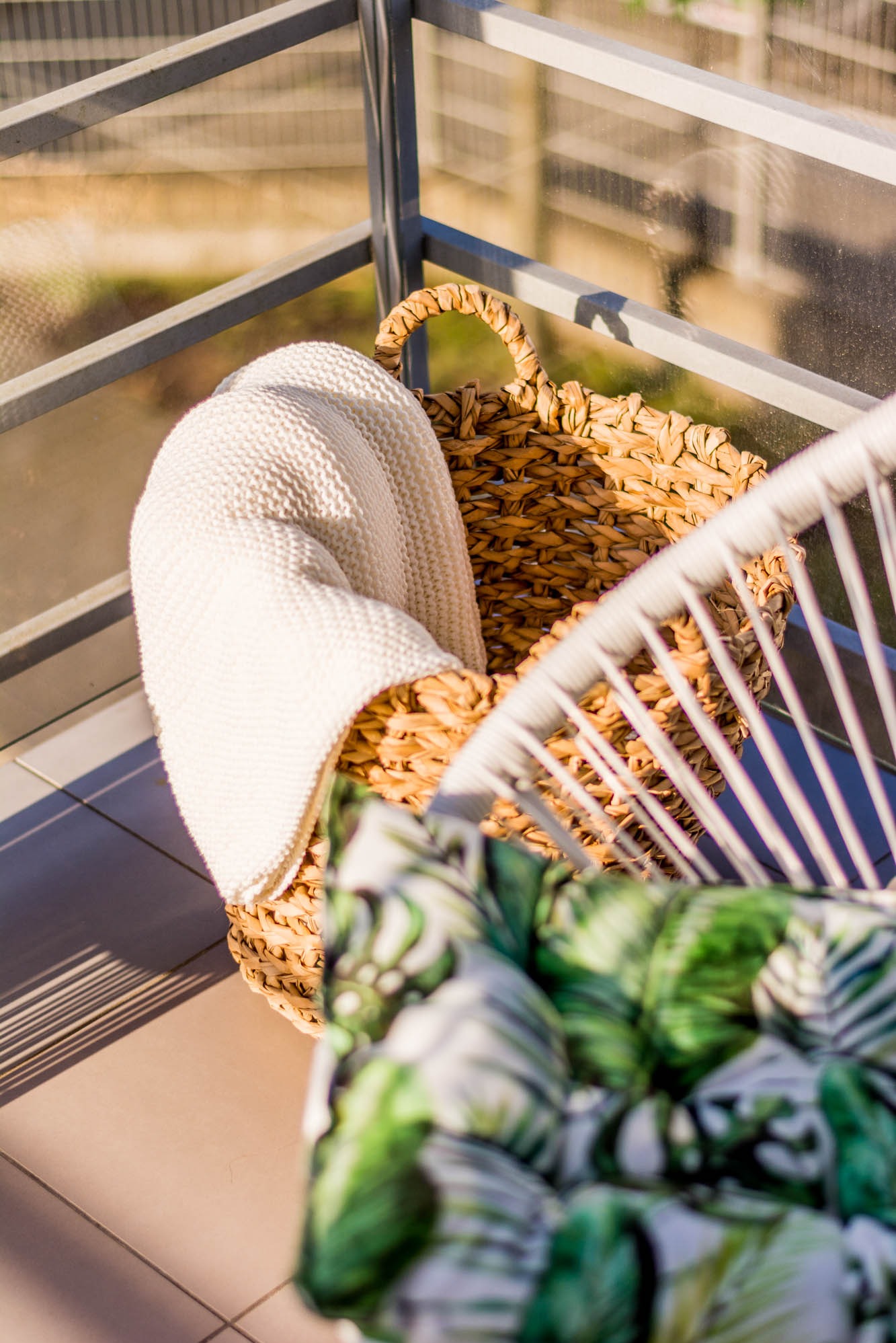 Słoneczny balkon: wiklinowy kosz z kremowym pledem obok białego krzesła z poduszką w tropikalne liście, widok z góry na taras w ciepłym świetle.