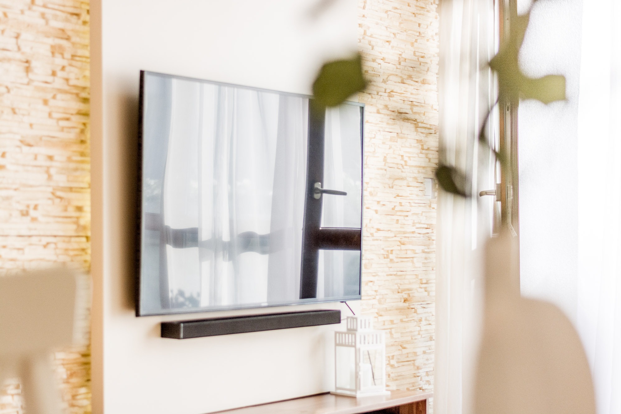 Telewizor na ścianie z kamienia dekoracyjnego, pod nim soundbar i lampion. W odbiciu widać okno z zasłoną. Fragment rośliny w kadrze.