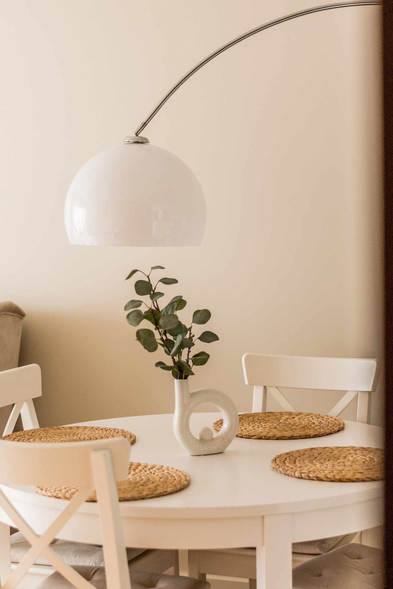 Jasne wnętrze jadalni: okrągły stół z białym blatem, rattanowe podkładki, designerski wazon z zielonymi gałązkami pod lampą z półokrągłym kloszem.