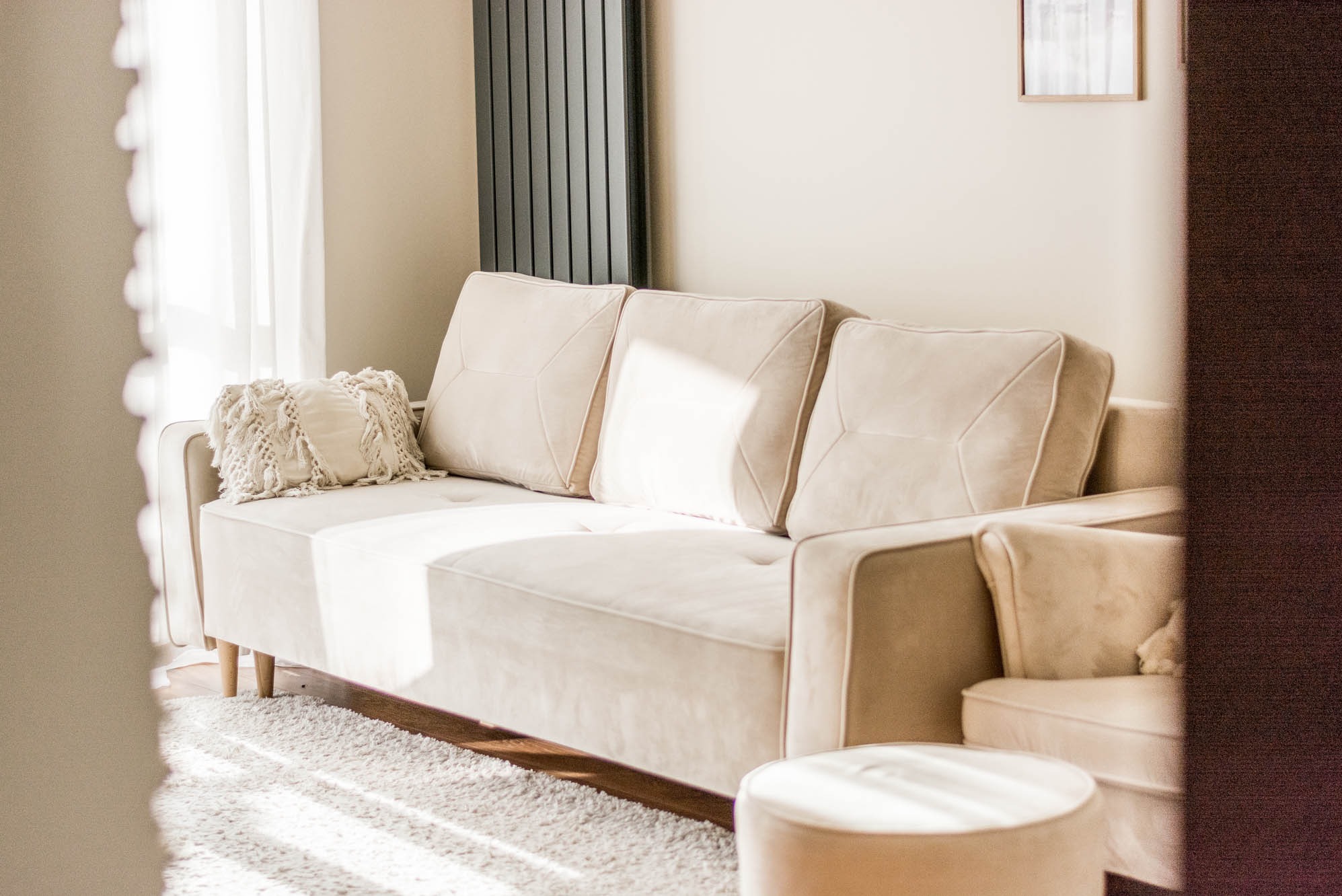 Jasny salon z beżową sofą, poduszkami i pufą. Minimalistyczny design, miękki dywan. Lekkie, naturalne światło przez okno z firanką. Widok zza framugi drzwi.