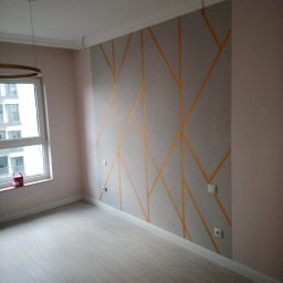 Rat-Bau Janusz Ratkiewicz - Pokój po malowaniu: szara ściana z geometrycznym wzorem z żółtych linii, jasne panele podłogowe, okno z widokiem na budynek. W tle widać wystające kable.