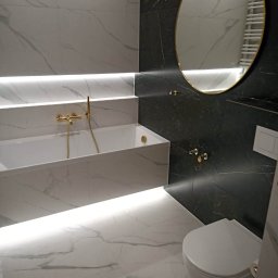 Rat-Bau Janusz Ratkiewicz - Elegancka łazienka z marmurowymi płytkami, podświetlaną wanną i złotymi akcentami, w tym okrągłe lustro i bateria. Nowoczesny design.