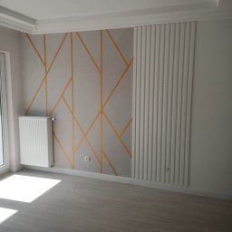 Rat-Bau Janusz Ratkiewicz - Wykończone wnętrze pokoju z dekoracyjną ścianą w geometryczne wzory i panelem ściennym. Widoczny grzejnik i jasna podłoga. Nowoczesny design.
