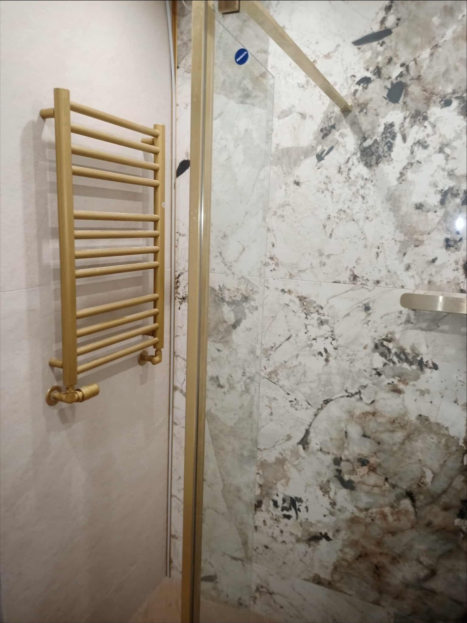 Złoty grzejnik drabinkowy na tle marmurowej ściany w łazience, obok kabiny prysznicowej ze złotymi ramami. Widok na wykończoną łazienkę z bliska.