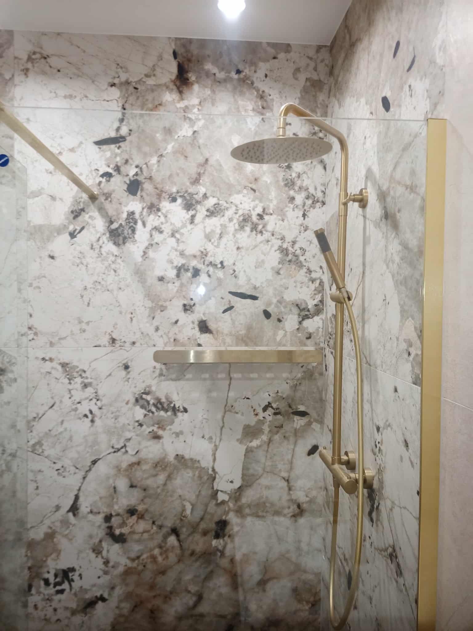 Wykończona łazienka z kabiną prysznicową typu walk-in, złota armatura i półka na tle płytek imitujących marmur.