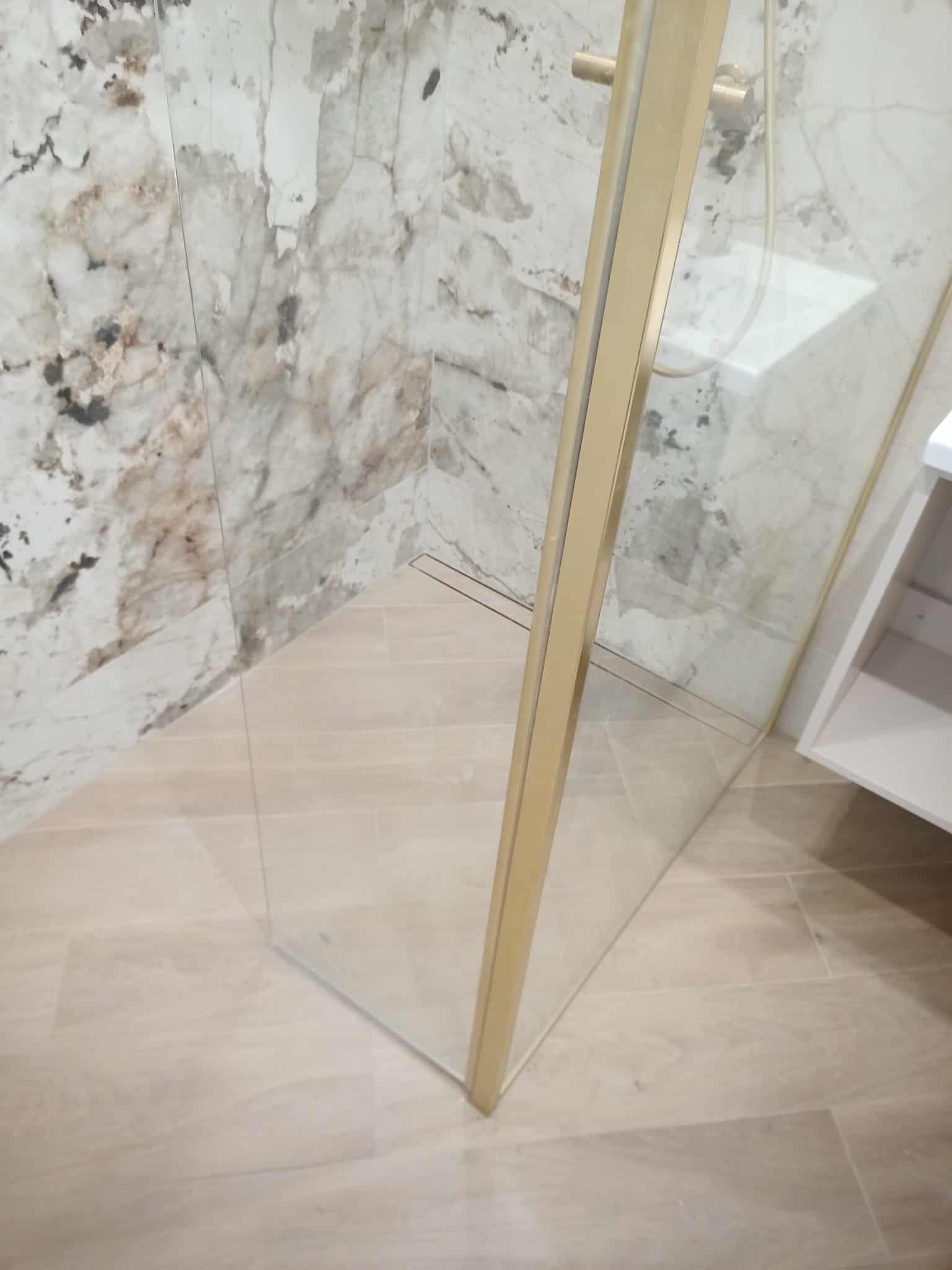 Nowoczesna kabina prysznicowa z złotymi profilami i marmurowymi płytkami na ścianach, w wykończonej łazience. Widoczne odpływ liniowy i fragment szafki łazienkowej.