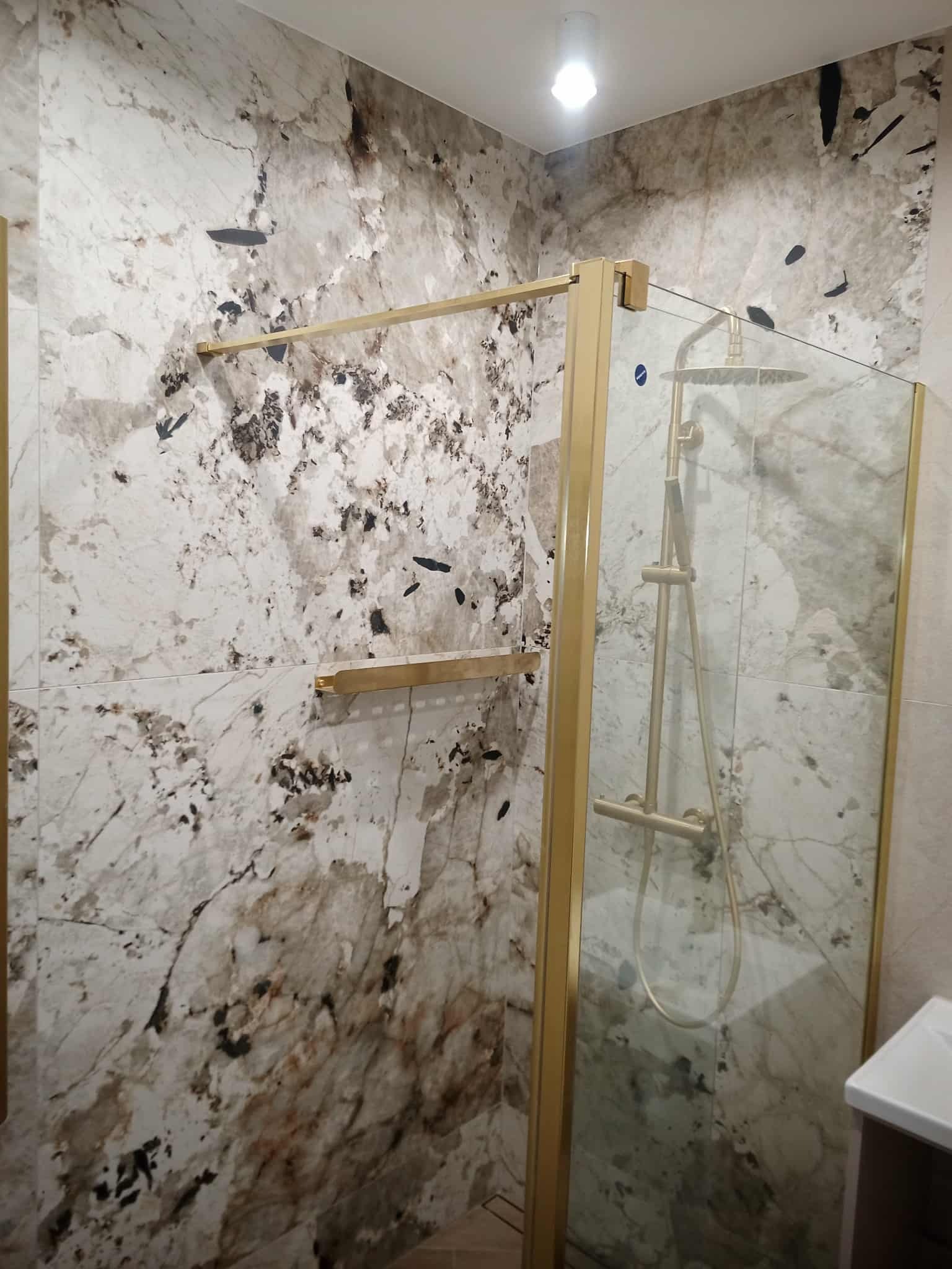 Wykończona łazienka z kabiną prysznicową w złotej ramie i ścianami wyłożonymi płytkami imitującymi marmur z ciemnymi żyłami. Nowoczesny design.