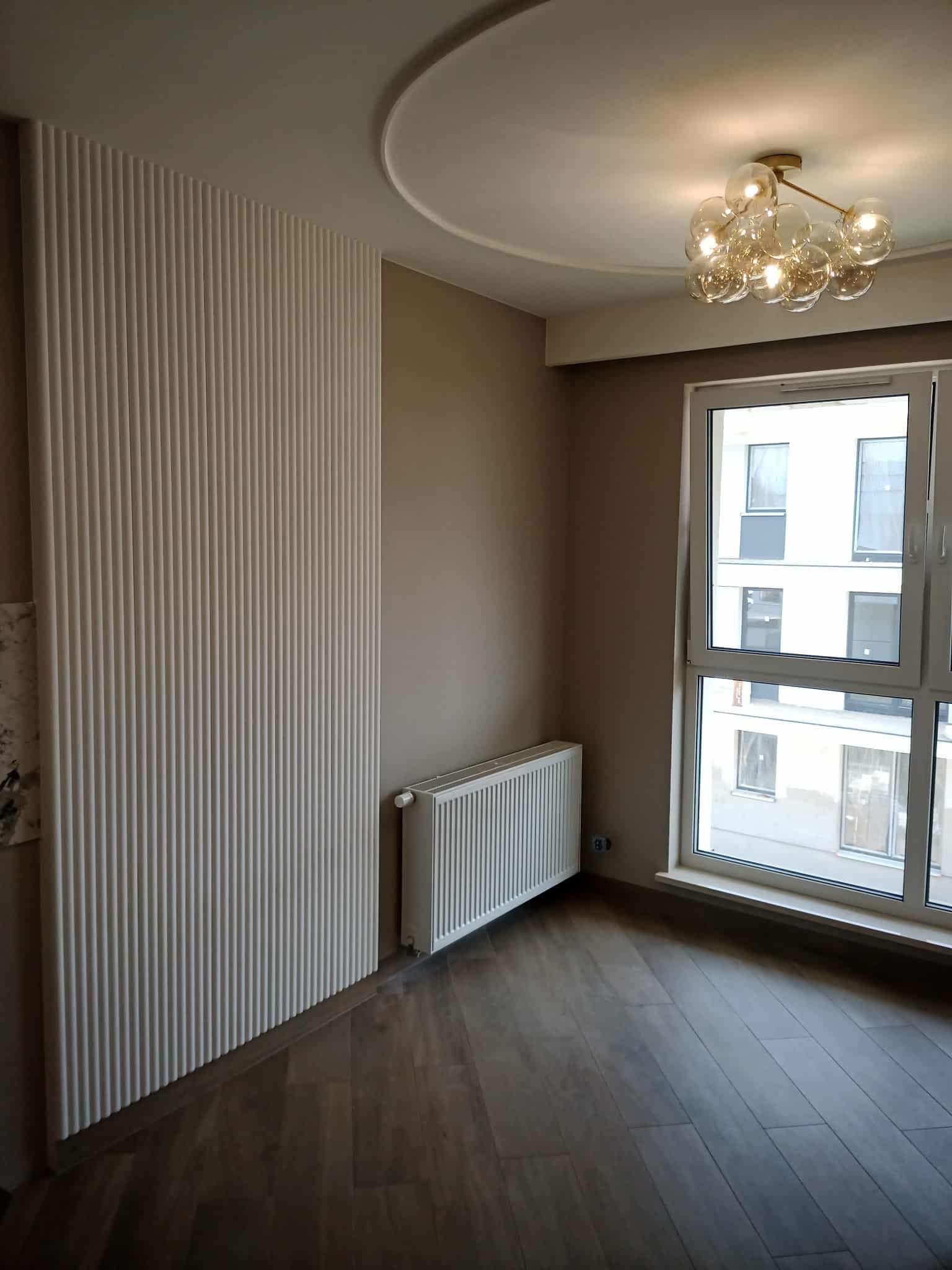 Nowoczesne wykończenie wnętrza: panel ścienny z lamelami, grzejnik, okno z widokiem na budynek i parkiet. Dekoracyjna lampa sufitowa.