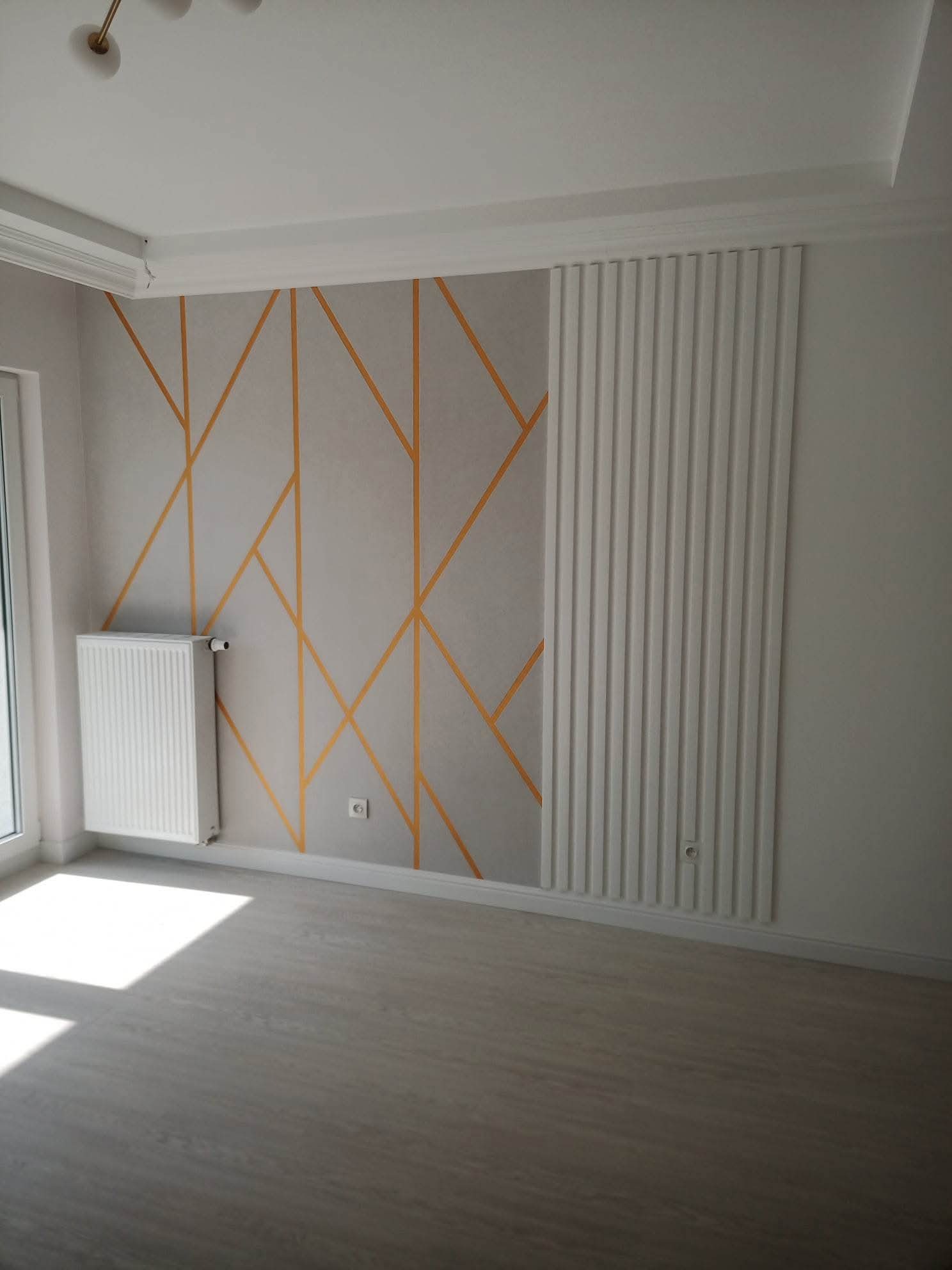 Wykończone wnętrze pokoju z dekoracyjną ścianą w geometryczne wzory i panelem ściennym. Widoczny grzejnik i jasna podłoga. Nowoczesny design.