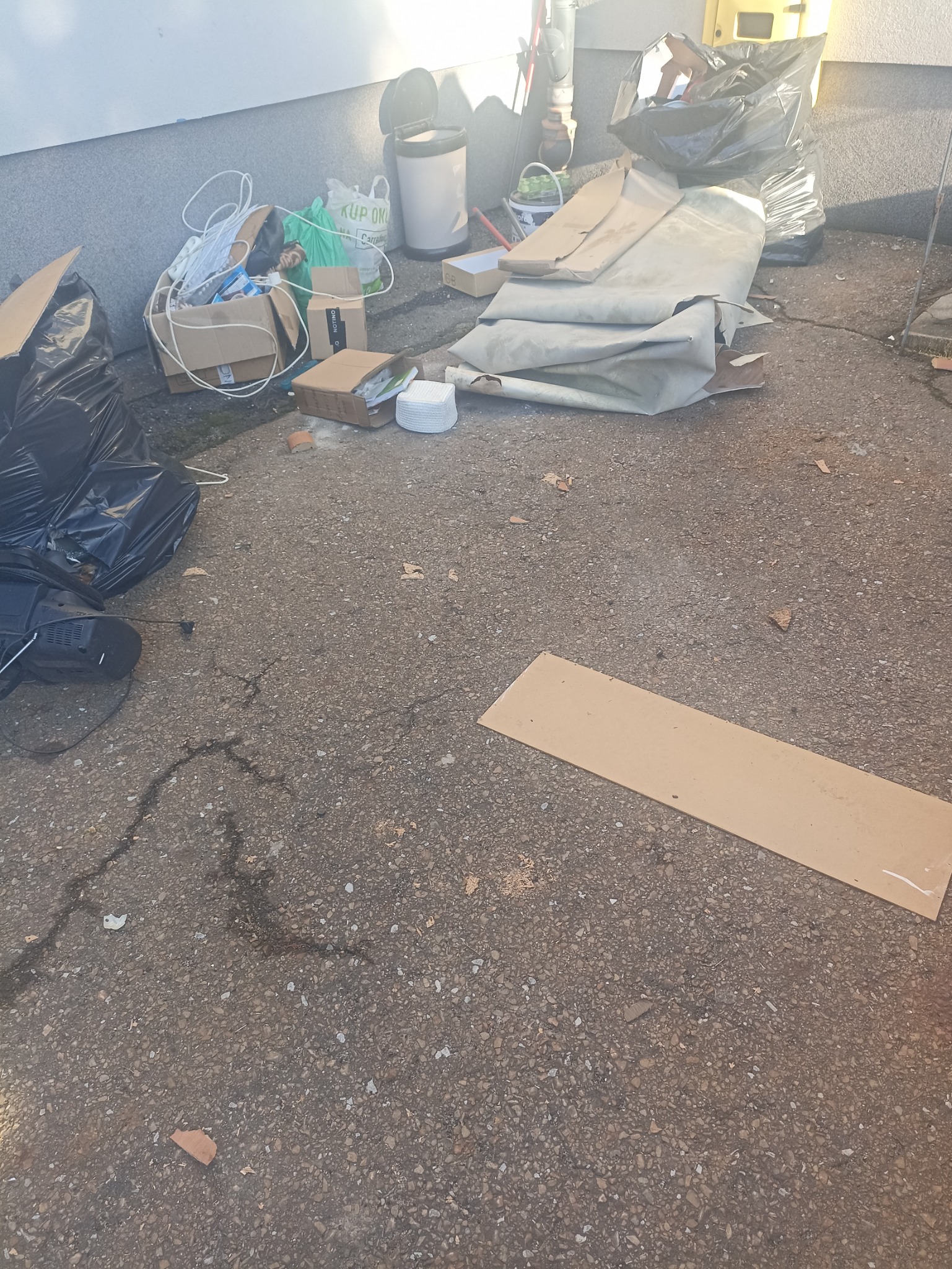 Sterta odpadów budowlanych i domowych na betonowym podłożu: kartony, worki na śmieci, resztki materiałów. Widoczne ślady po zalaniu.