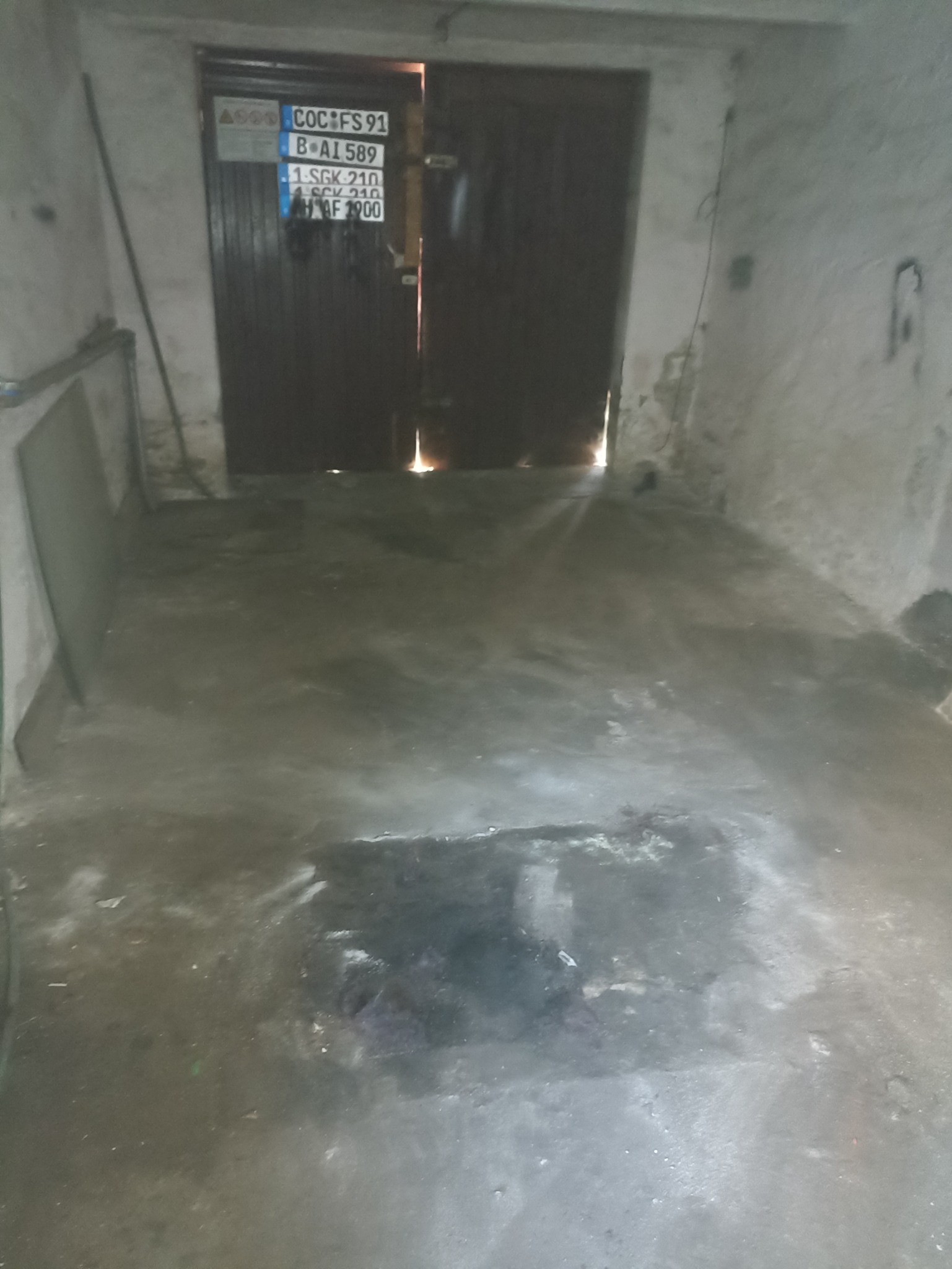 Opróżnione pomieszczenie z betonową posadzką, widoczne otwarte, ciemne drzwi z tablicami rejestracyjnymi i ślady na ścianach. Ponure oświetlenie.