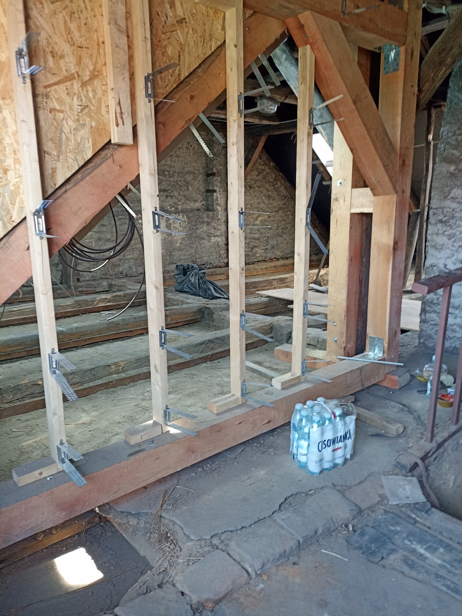 Konstrukcja szkieletowa poddasza z drewnianych belek, metalowych łączników i butelką wody mineralnej w tle, ukazująca etap prac budowlanych.