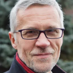 Portret mężczyzny w okularach z szpakowatą brodą, w ciemnej marynarce z czerwoną podszewką na tle zieleni. Uśmiech, zmarszczki, naturalne światło.