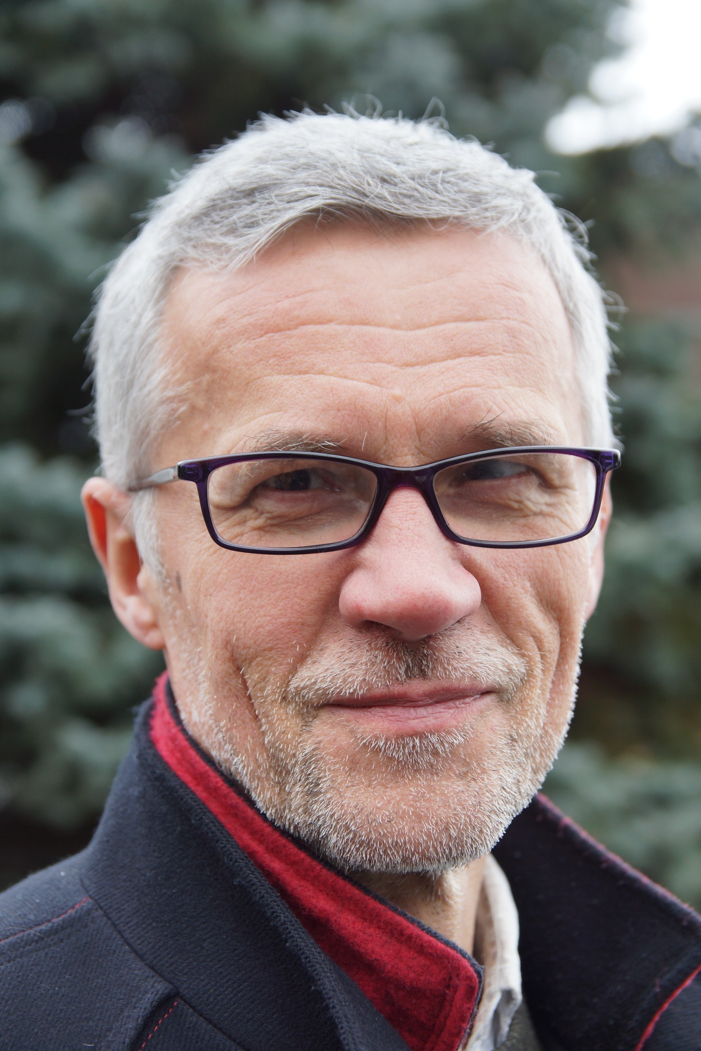 Portret mężczyzny w okularach z szpakowatą brodą, w ciemnej marynarce z czerwoną podszewką na tle zieleni. Uśmiech, zmarszczki, naturalne światło.