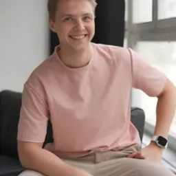 Uśmiechnięty blondyn siedzi na czarnej sofie, ubrany w różową koszulkę i beżowe spodnie. W tle okno i czarna zasłona. Widoczny zegarek na nadgarstku.