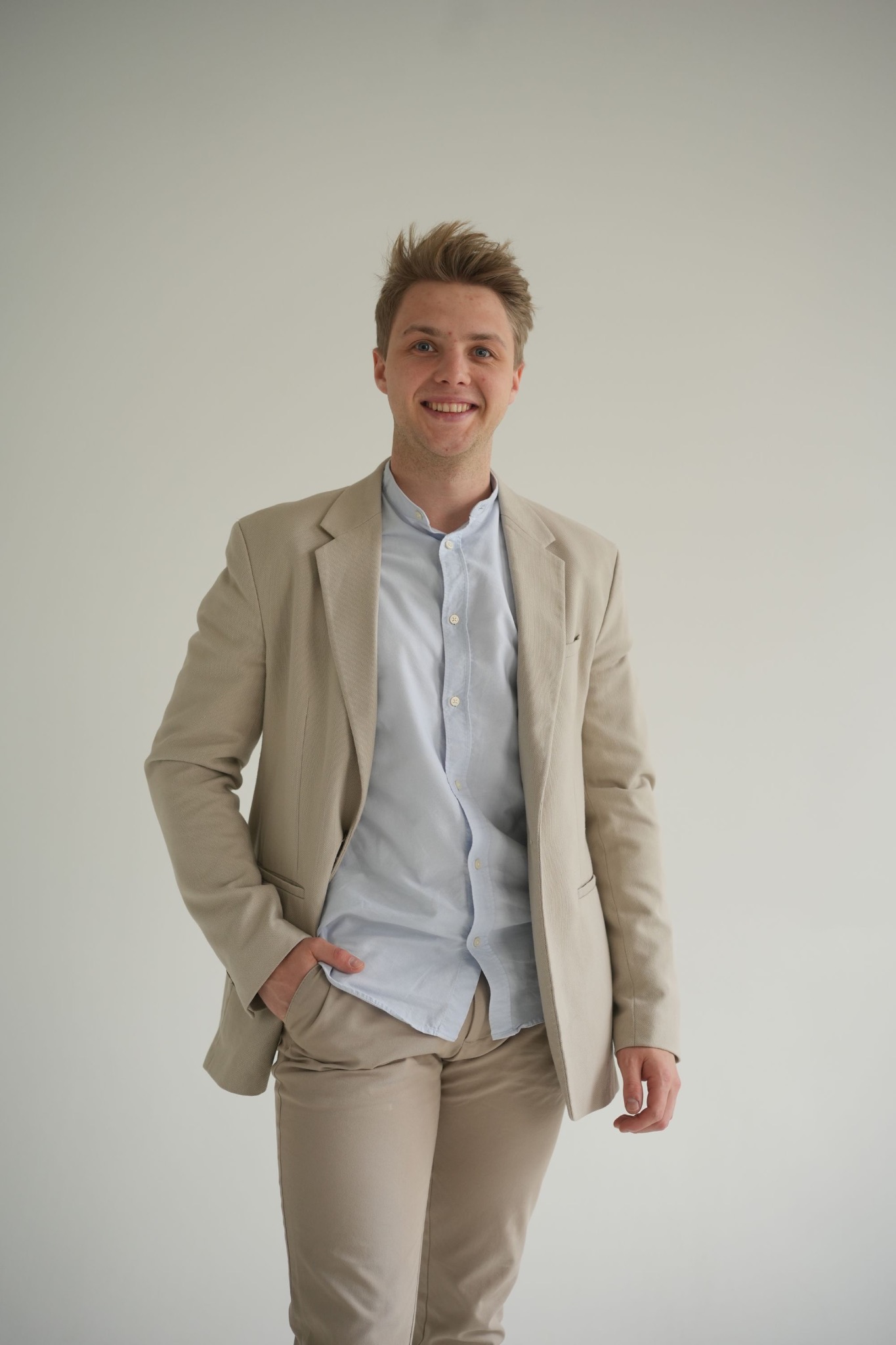Uśmiechnięty mężczyzna w beżowym garniturze i błękitnej koszuli, pozujący na jasnym tle. Ręka w kieszeni, swobodny styl, profesjonalny wygląd.