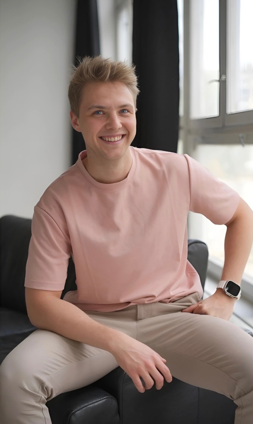 Uśmiechnięty blondyn siedzi na czarnej sofie, ubrany w różową koszulkę i beżowe spodnie. W tle okno i czarna zasłona. Widoczny zegarek na nadgarstku.