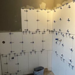 Gukasov.ru - Białe płytki ułożone na ścianie w łazience, z niebieskimi krzyżykami dystansowymi. Widoczny fragment podłogi i ściana w surowym stanie.