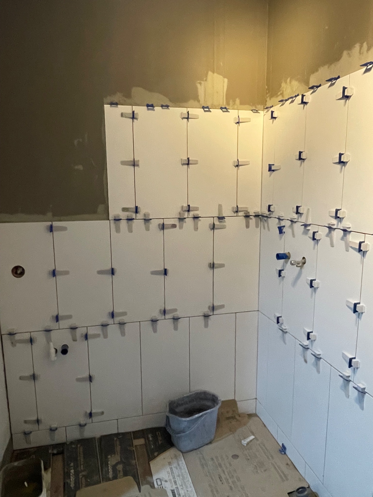 Białe płytki ułożone na ścianie w łazience, z niebieskimi krzyżykami dystansowymi. Widoczny fragment podłogi i ściana w surowym stanie.