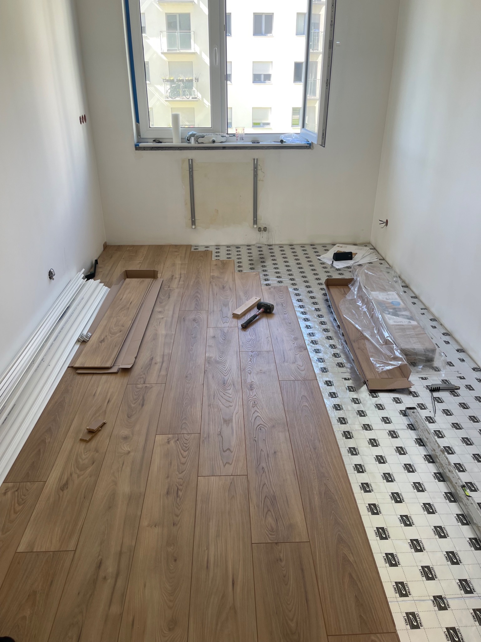 Układanie paneli podłogowych w jasnym pokoju z widokiem na budynek. Częściowo ułożone panele, narzędzia i podkład na podłodze. Etap prac wykończeniowych.