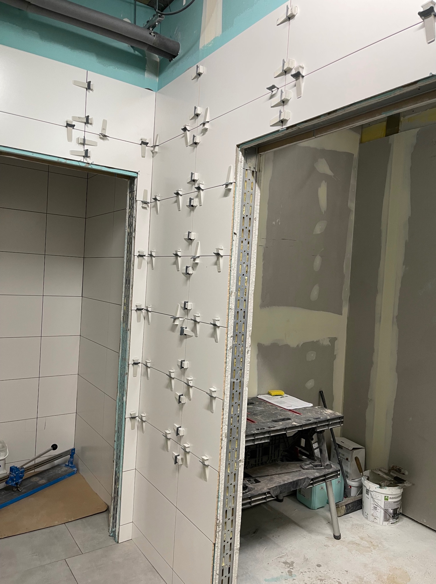 Białe płytki ścienne z plastikowymi krzyżykami dystansowymi w trakcie układania. Widoczne narzędzia i fragmenty ścian przygotowanych do dalszych prac wykończeniowych.