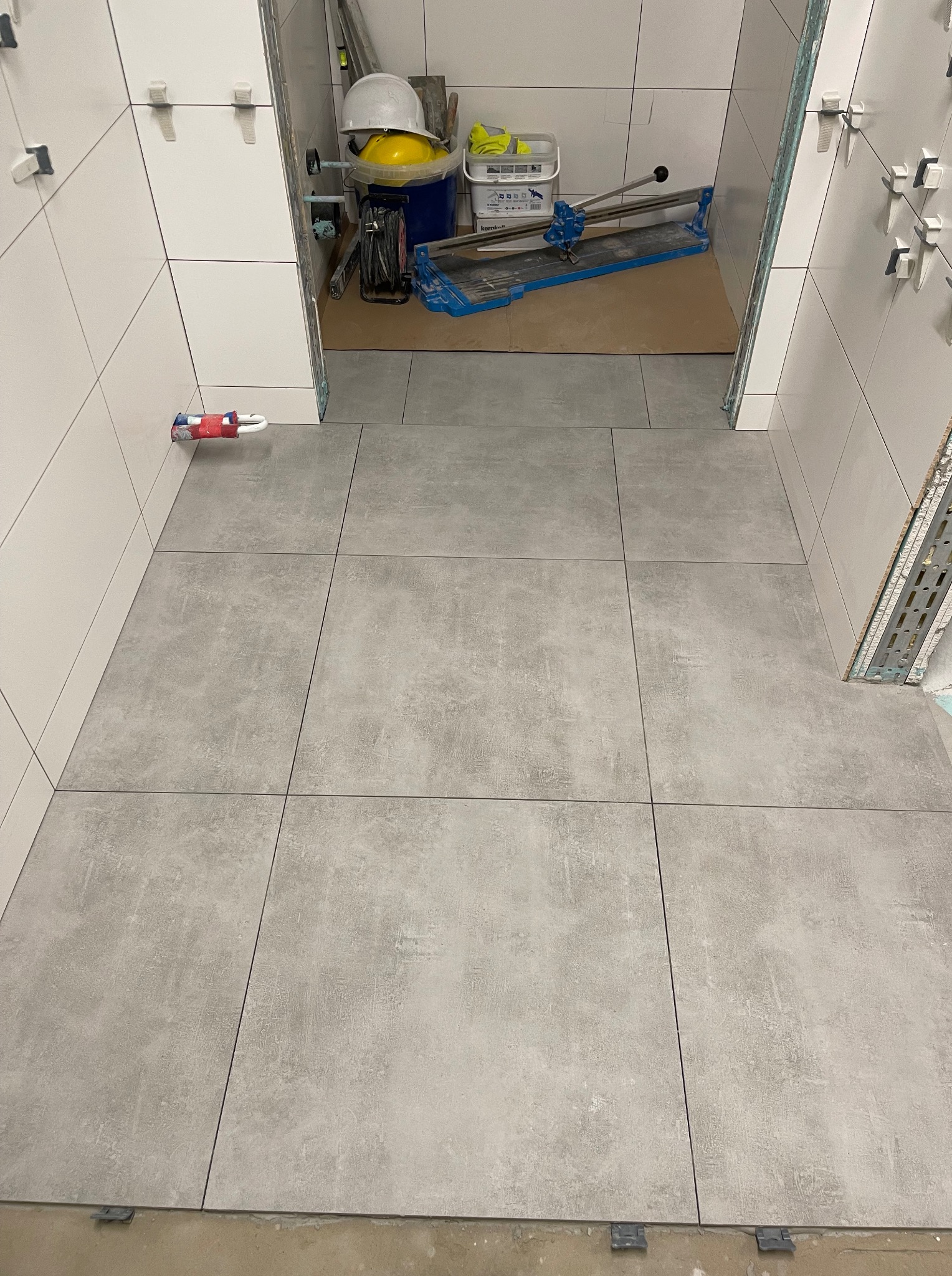 Ułożone szare płytki podłogowe w łazience, widoczne narzędzia i akcesoria glazurnicze, białe płytki ścienne w trakcie montażu, elementy instalacji wodnej.