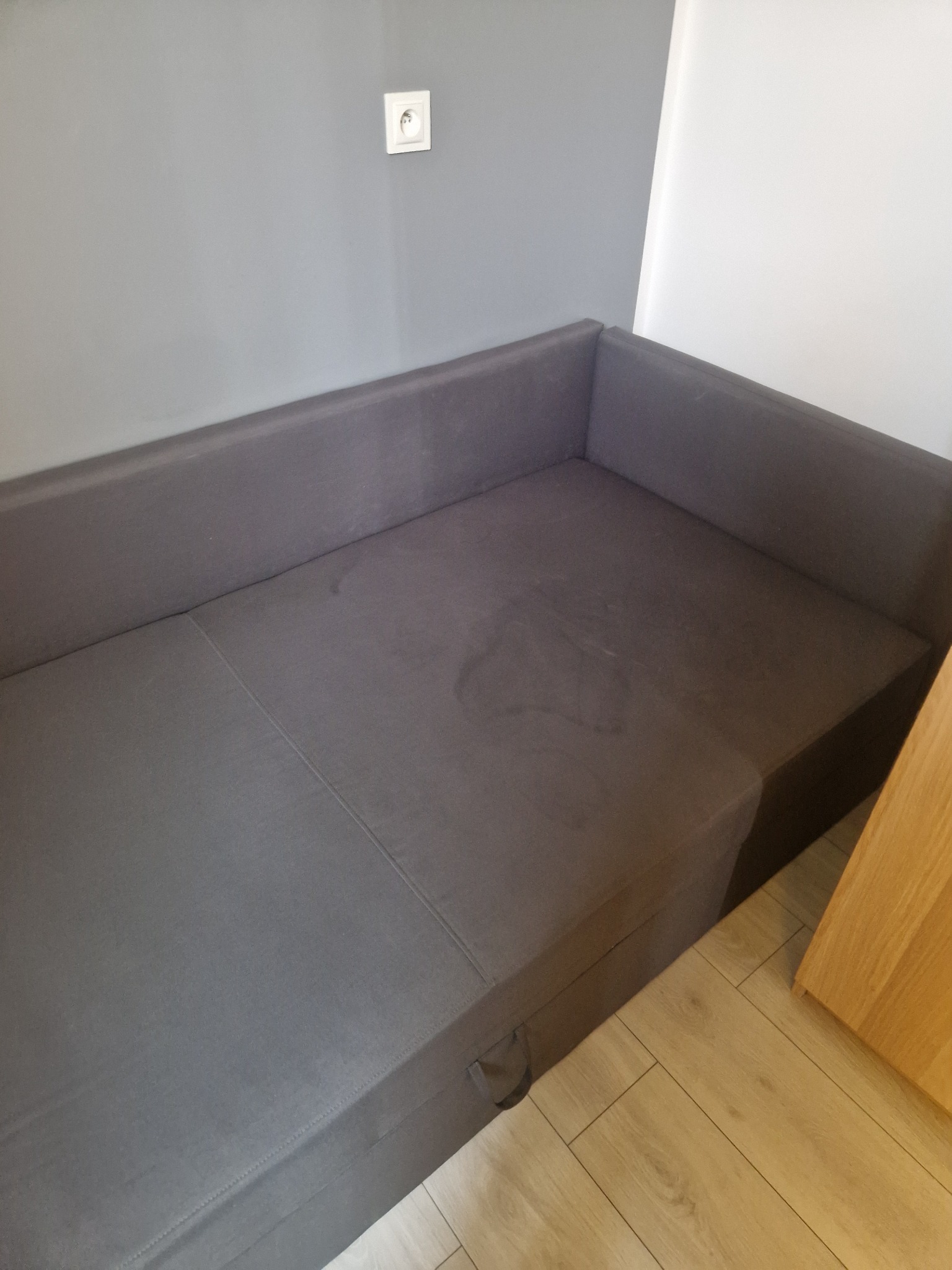 Szara, rozłożona kanapa z plamami na siedzisku, widoczna w rogu pomieszczenia z jasną podłogą i gniazdkiem na szarej ścianie. Widoczny uchwyt szuflady pod siedziskiem.