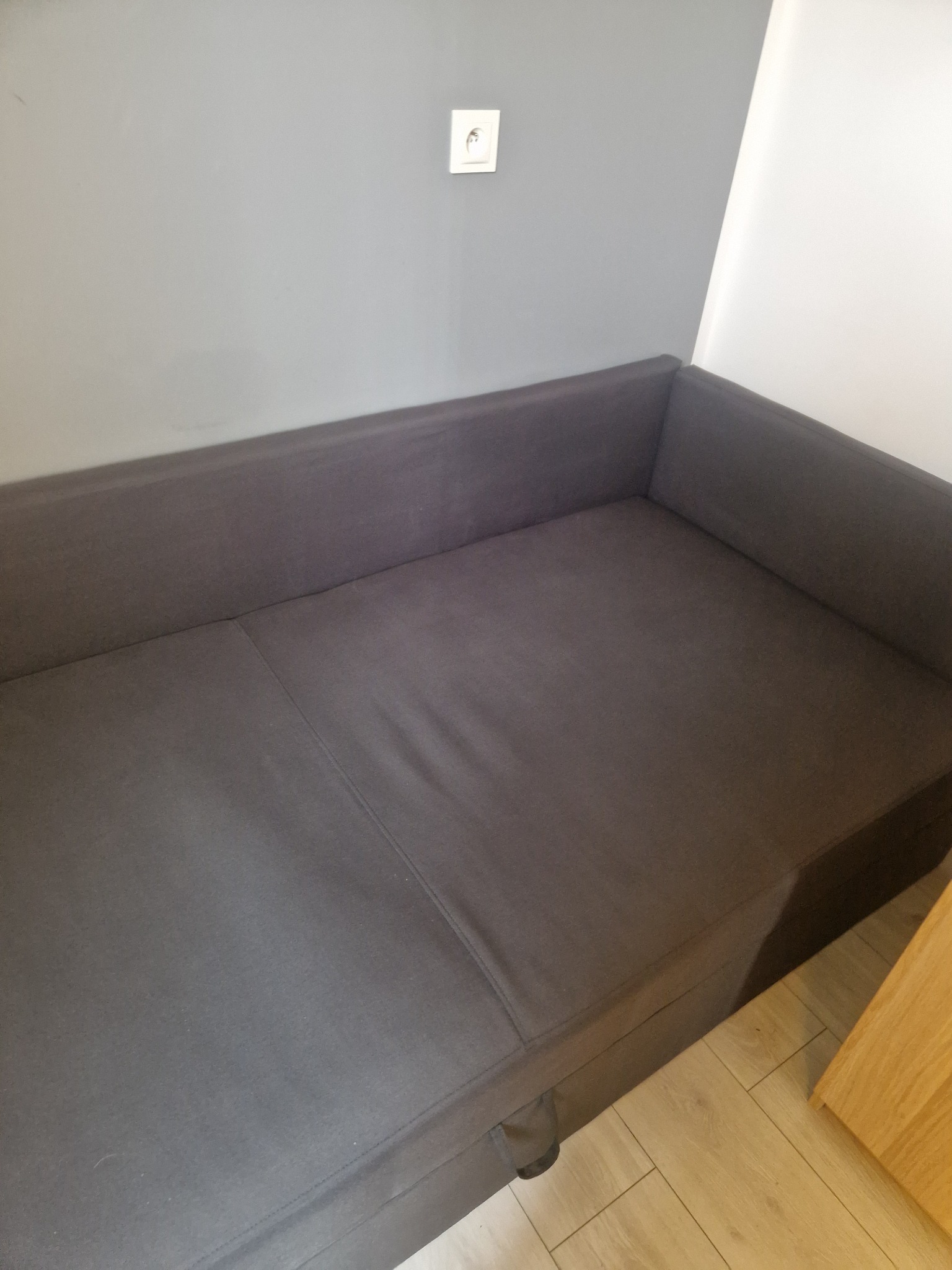 Szara, rozłożona kanapa w rogu pokoju, widoczny fragment drewnianej podłogi i kąt ściany. Minimalistyczne wnętrze, proste linie, stonowana kolorystyka.