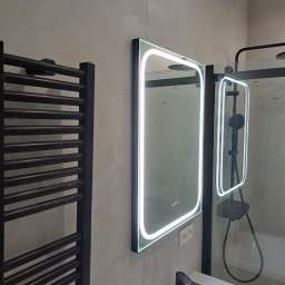 Artrewnetrza - Nowoczesna łazienka z podświetlanym lustrem, czarną armaturą i grzejnikiem drabinkowym. Minimalistyczny design, jasne płytki, kabina prysznicowa w tle.