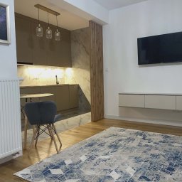 Artrewnetrza - Nowoczesne wnętrze: beżowa kuchnia z marmurowym blatem, drewniana ścianka działowa, TV na ścianie, dywan w geometryczne wzory, krzesło i grzejnik.