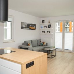 Artrewnetrza - Jasne wnętrze salonu z aneksem kuchennym: szara sofa, drewniany parkiet, białe ściany, okno z roletami plisowanymi i wyspa kuchenna z płytą indukcyjną.