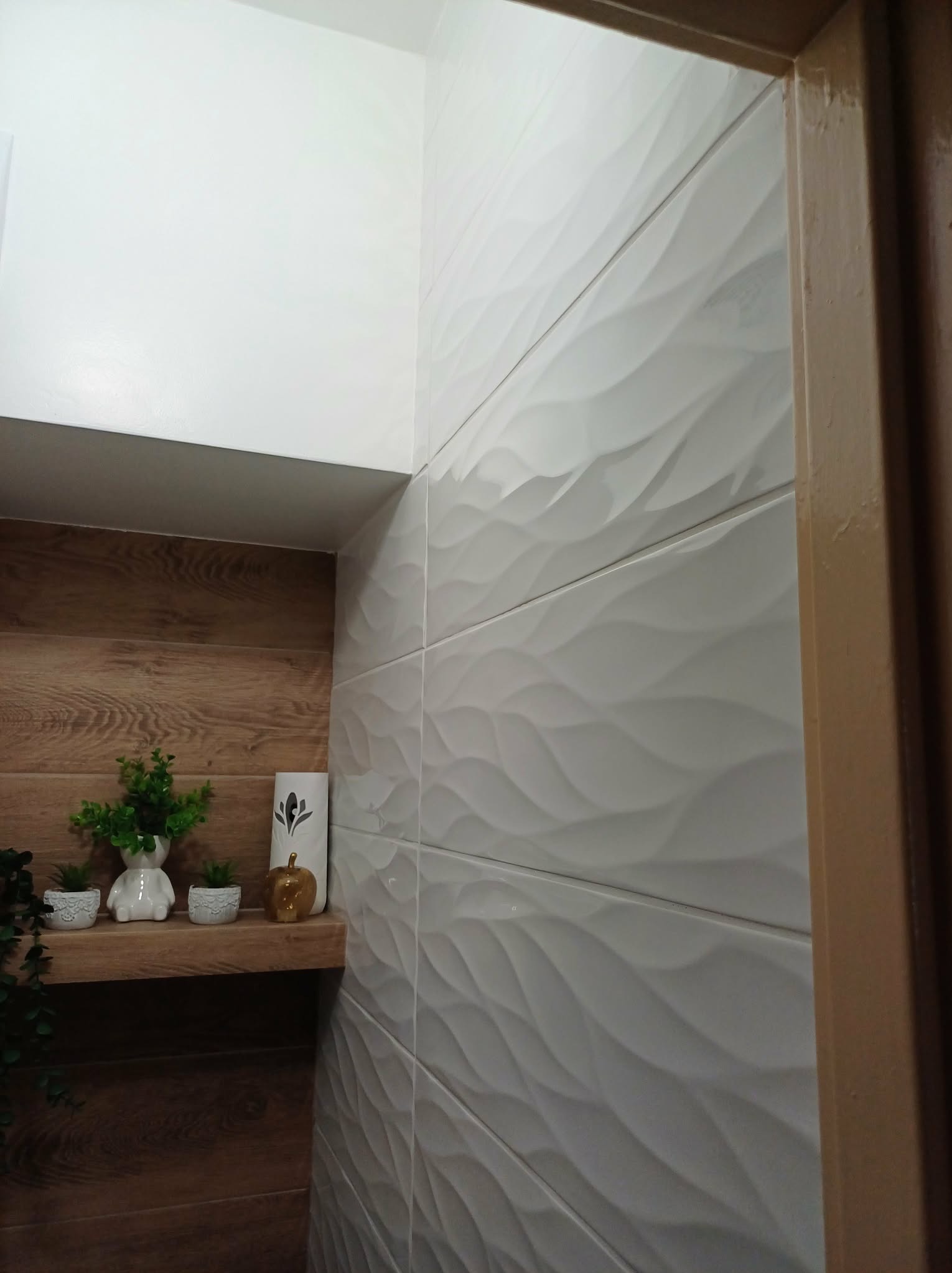 Białe, faliste płytki ścienne w łazience, drewniana półka z dekoracjami, ciepłe oświetlenie. Detal wykończenia wnętrza z bliska.