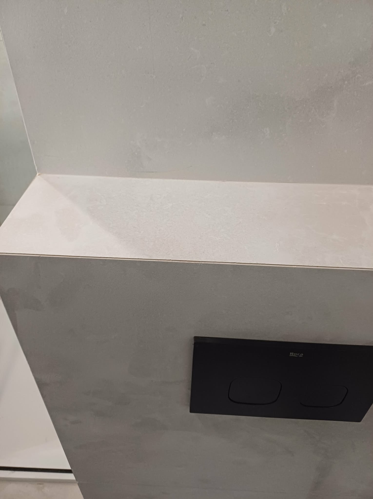 Minimalistyczna łazienka: szare płytki imitujące beton, czarny panel spłukujący Roca. Nowoczesne wykończenie, stonowana kolorystyka, surowy design.