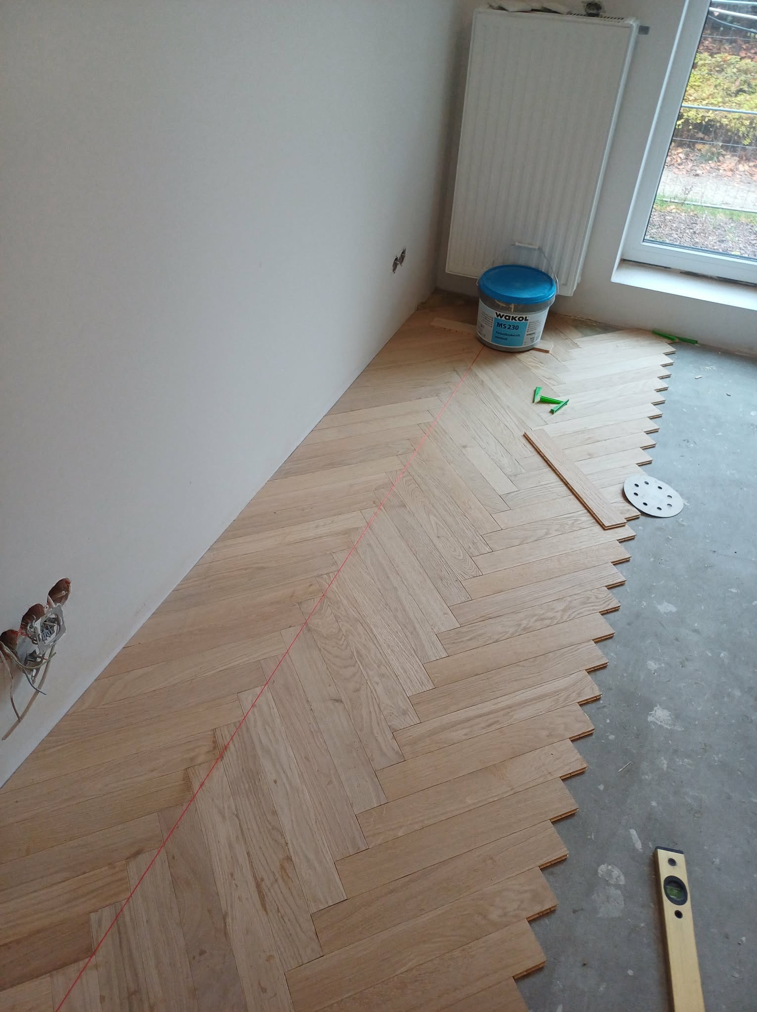 Układanie parkietu w jodełkę, widoczny laser wyznaczający linię, klej Wakol, poziomica i tarcza szlifierska. Częściowo ułożona podłoga z drewnianych desek.