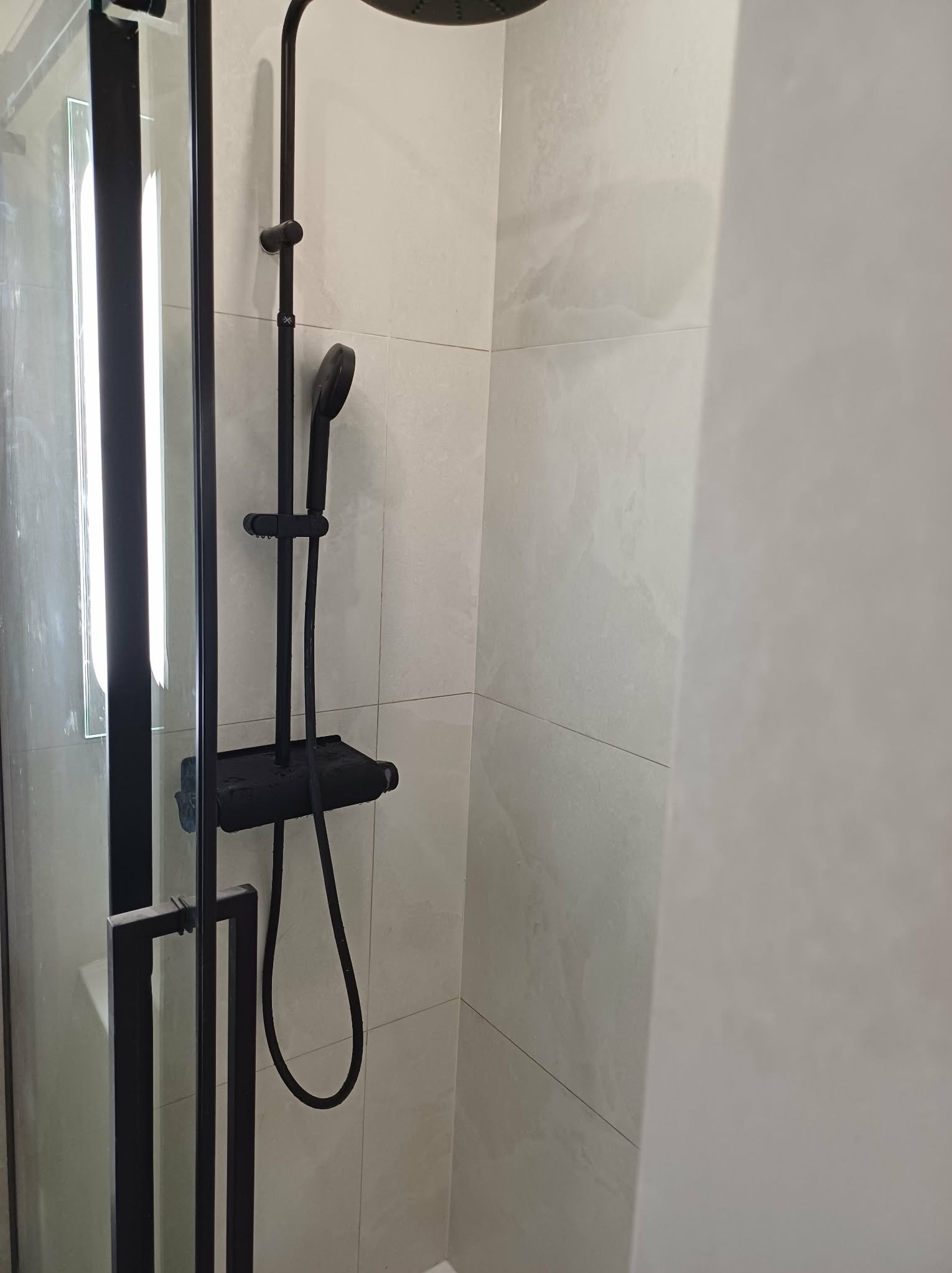 Kabina prysznicowa z czarną armaturą na tle jasnych, marmurkowych płytek. Minimalistyczny design, nowoczesne wykończenie łazienki. Widoczny fragment szklanej ściany.