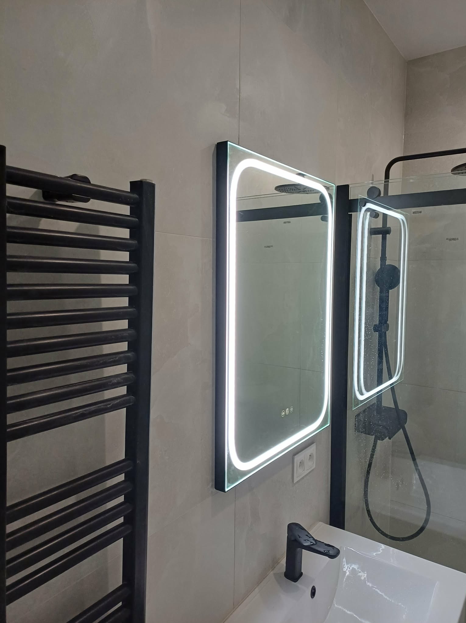 Nowoczesna łazienka z podświetlanym lustrem, czarną armaturą i grzejnikiem drabinkowym. Minimalistyczny design, jasne płytki, kabina prysznicowa w tle.