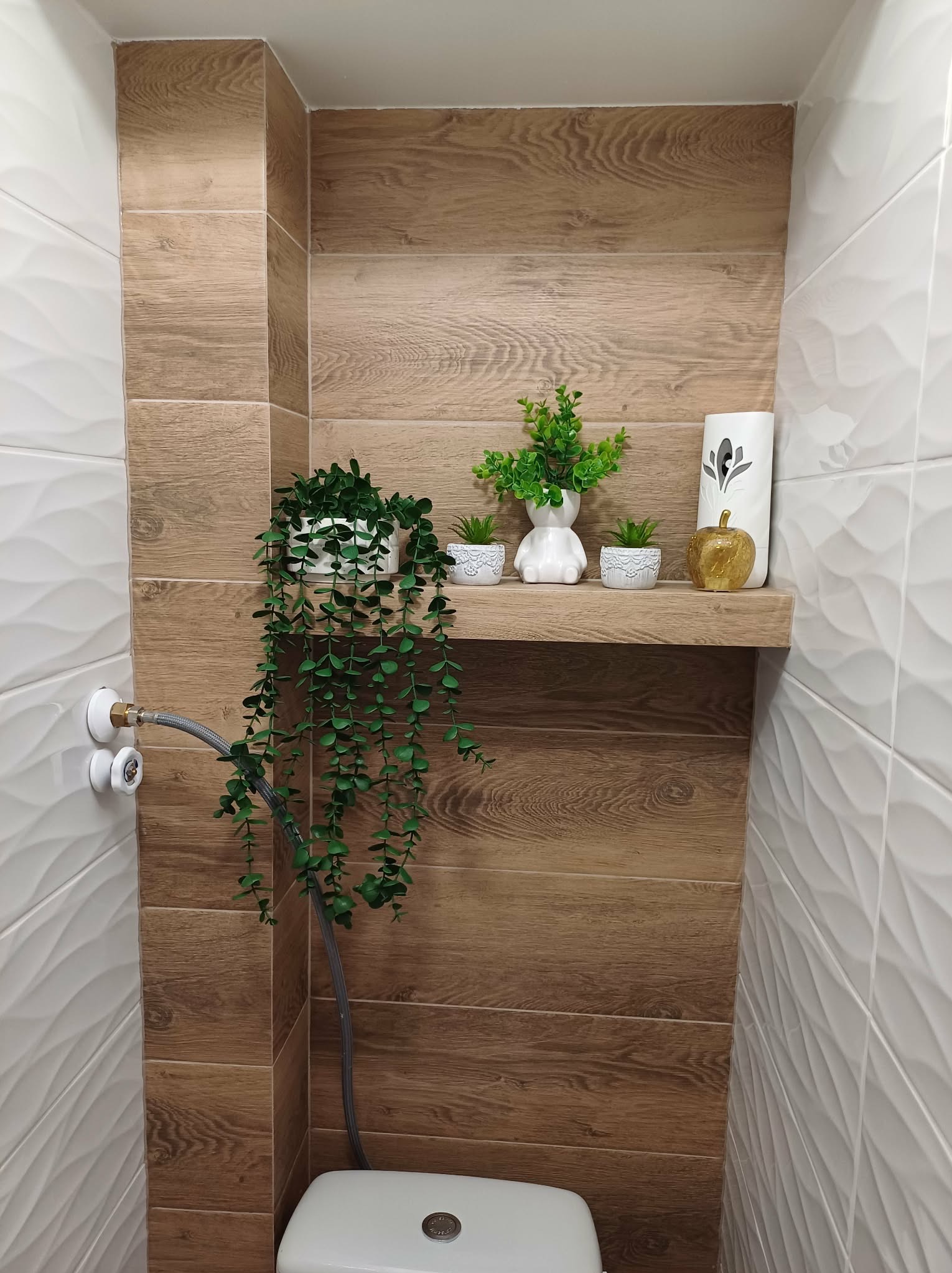 Nowoczesna łazienka z drewnopodobnymi płytkami i dekoracyjną półką z roślinami. Białe, faliste płytki na ścianie kontrastują z ciepłym odcieniem drewna.