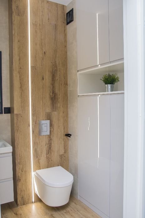 Nowoczesna łazienka z podwieszaną toaletą, drewnopodobnymi płytkami z oświetleniem LED i białymi szafkami z rośliną doniczkową. Minimalistyczny design.